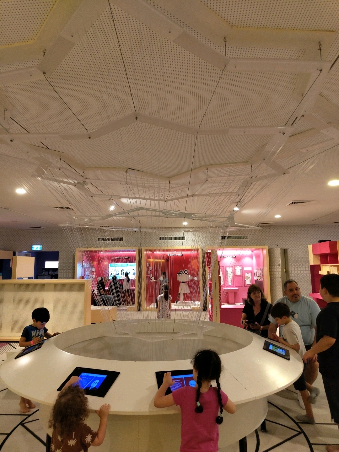 The image depicts an indoor exhibition space, likely a museum or interactive learning center, with a focus on technology and media. The foreground features a large, circular interactive table with embedded screens, surrounded by several children. To the left, a young boy wearing a blue t-shirt with the word "MINI" visible on it is looking at a screen. In front of the table, two girls are engaged with the interactive displays; one in a pink t-shirt with braided hair, and another with curly hair facing away from the camera. In the background, there is a family unit consisting of a man, a woman, and a boy, observing the exhibit. Behind them, display cases are visible, showcasing various artifacts and themes such as "MUSIC," "CINEMA," and "FASHION." The overall environment is well-lit with modern architecture, including a perforated ceiling with intricate wire installations. The scene is consistent with the exhibits found at the Bloomfield Science Museum in Jerusalem, though the question asks for Tel-Aviv. Without further context, a definitive location in Tel-Aviv cannot be confirmed from the image alone.