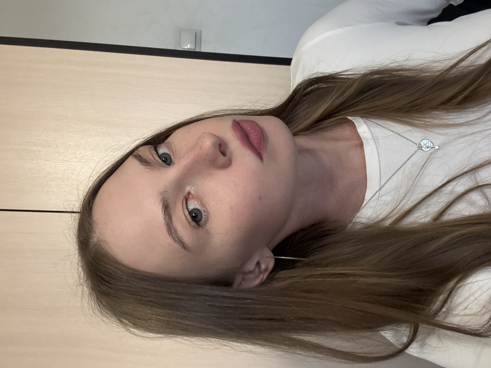 A young woman with long, light brown hair and light green eyes is photographed from the chest up, with her head tilted to her left. She is wearing a white top and a silver necklace with a heart-shaped pendant and a small key. Her lips are painted with a matte pink lipstick. Her left ear is visible, adorned with a long, thin silver earring. The background appears to be an interior wall, possibly in an apartment or office building, with vertical wooden paneling on the left and a plain wall on the right. A dark vertical line suggests a doorway or seam. The lighting is soft, originating from the front and slightly from the left. The image is a selfie, likely taken indoors. The location is not specified in the image and cannot be determined from the visual information alone. There are no indications of the city of Perm, Russia.