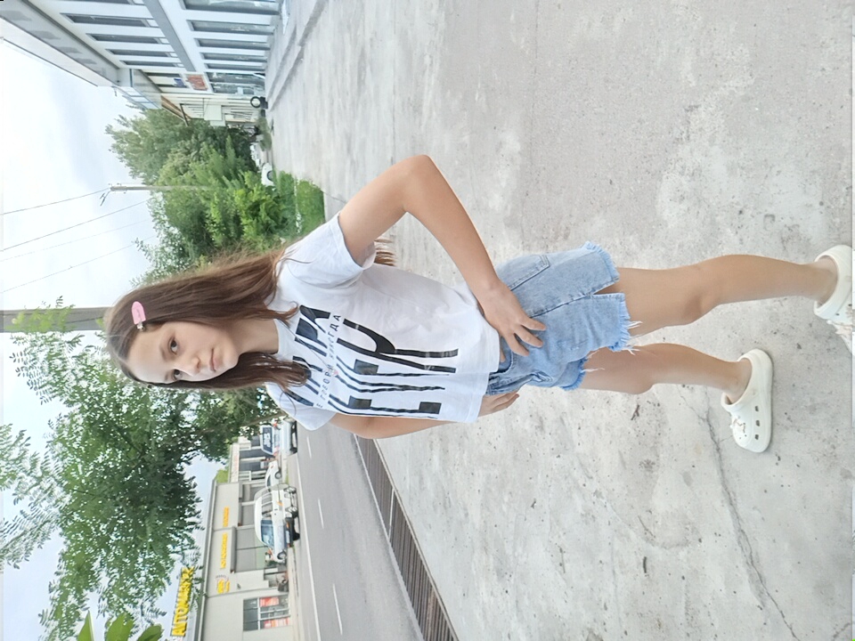 A young girl with long brown hair, wearing a white t-shirt with black lettering and light blue denim shorts, stands on a sidewalk in Angren, Uzbekistan. She has a pink hair clip in her hair and is wearing white clogs. The girl is looking directly at the camera with her hands on her hips. In the background, there is a street with cars, a building with shops, and trees. The sidewalk is made of concrete, and there is a curb and a road. The sky is overcast.