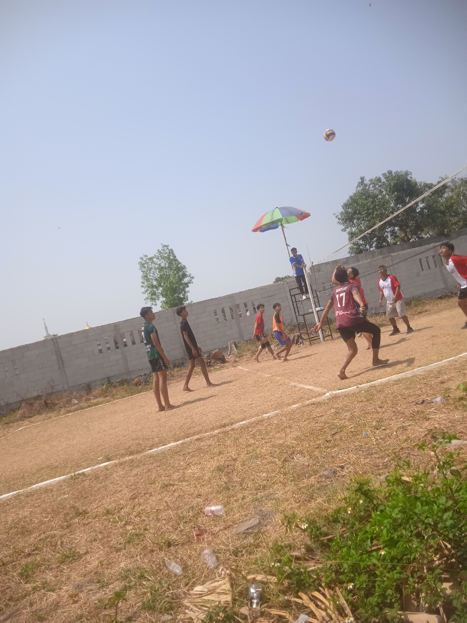 The image depicts a volleyball game taking place on a dirt court, likely located in Klodran, Indonesia, given the context provided. Several individuals, appearing to be young men, are actively engaged in the game. One player in a maroon jersey with the number "17" on it is in the foreground, mid-action, facing away from the camera. Other players are visible on both sides of the net, some in motion, suggesting the game is in progress. A referee or scorekeeper is positioned on a raised platform with an umbrella providing shade. The background shows a grey brick wall, trees, and a clear sky. The court surface is covered with dry grass and dirt. Scattered on the ground are discarded plastic bottles and debris, indicating a casual or informal setting. The overall scene captures a moment of athletic activity in an outdoor environment.