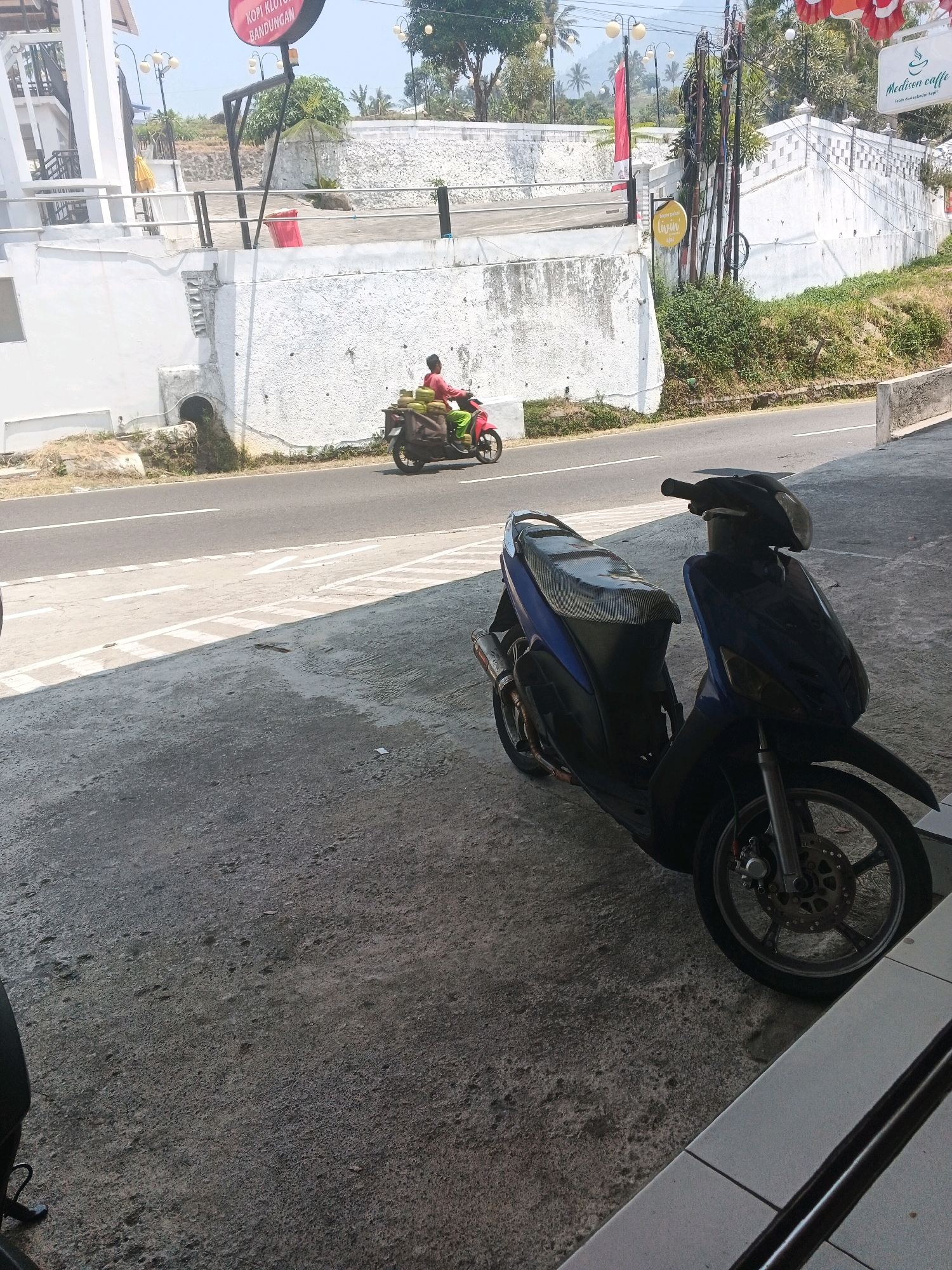 In Pakopen, Indonesia, a motorcycle rider wearing a red shirt and green pants is seen riding a motorcycle on a paved road. The motorcycle is loaded with green coconuts in a brown bag. In the foreground, a blue and black scooter is parked on a concrete surface. Behind the road, a white building with stone accents and a white wall with a textured finish are visible. Trees and power lines are also present in the background, with a clear sky overhead. A sign with red and white flags and a red circular sign with text are also visible.