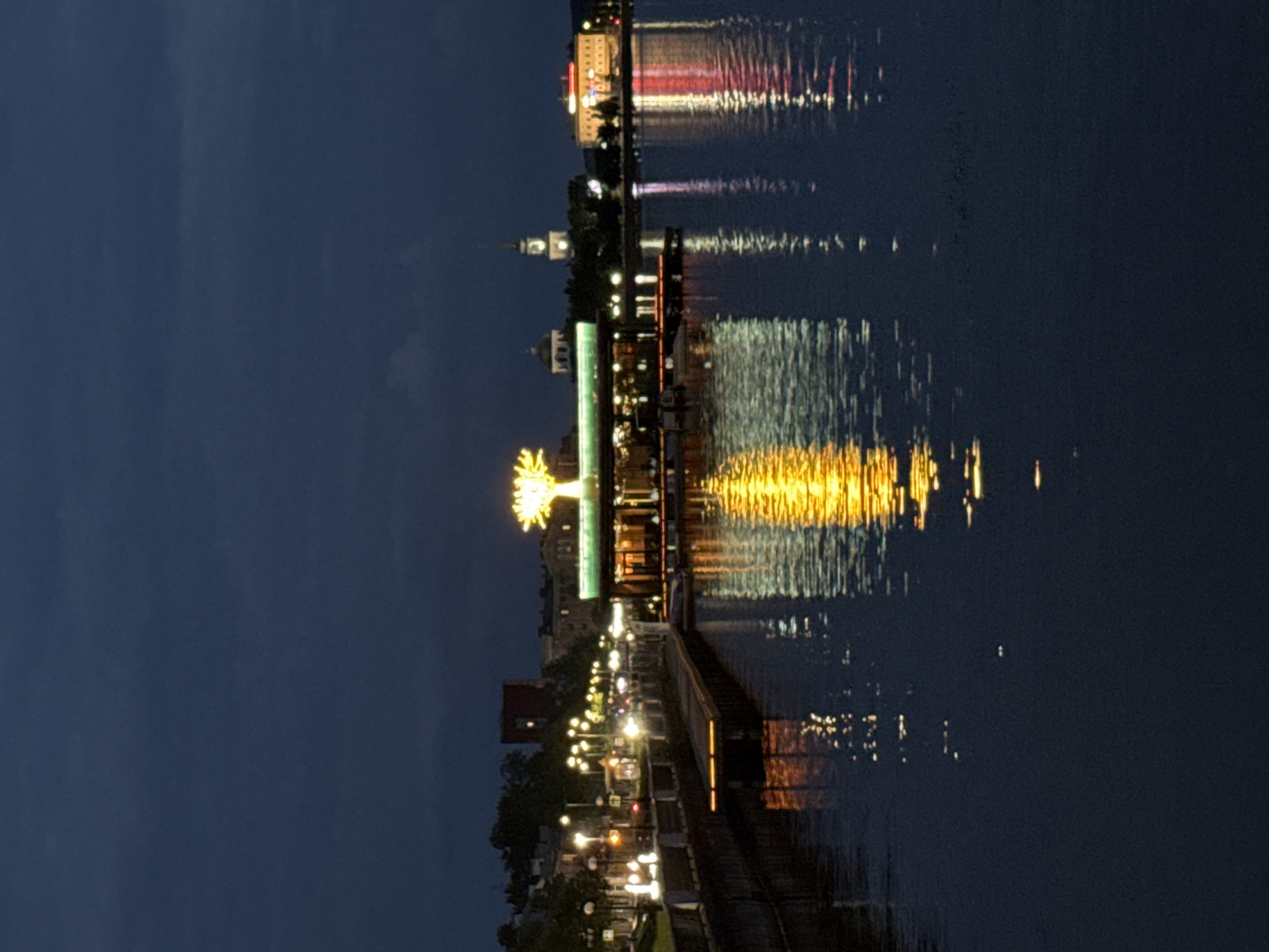 This is a night-time, vertical, medium-distance shot of a waterfront area in Votkinsk, Russia. The image captures a serene scene with artificial lights reflecting on the dark water.
The left side of the image shows a waterfront promenade lined with lampposts, casting a warm glow on the walkway. A low stone wall runs along the edge of the promenade. In the background, buildings of various sizes and architectural styles are visible, some with lit windows.
The center of the image features a modern building with a green roof, illuminated by a bright, stylized tree-shaped light installation. The reflections of this light, along with other lights from buildings across the water, create a shimmering pattern on the water's surface.
The right side of the image reveals a cityscape with a distinct church steeple, a domed building, and a larger, multi-story building with a red illuminated sign. The reflections of these structures and their lights are also visible on the water.
The water itself occupies the lower half of the image, dark and still, serving as a mirror for the lights of the city. The sky above is a deep, cloudy blue, indicating nighttime. There are no visible people in the frame. The overall atmosphere is tranquil and picturesque, highlighting the beauty of the city at night.