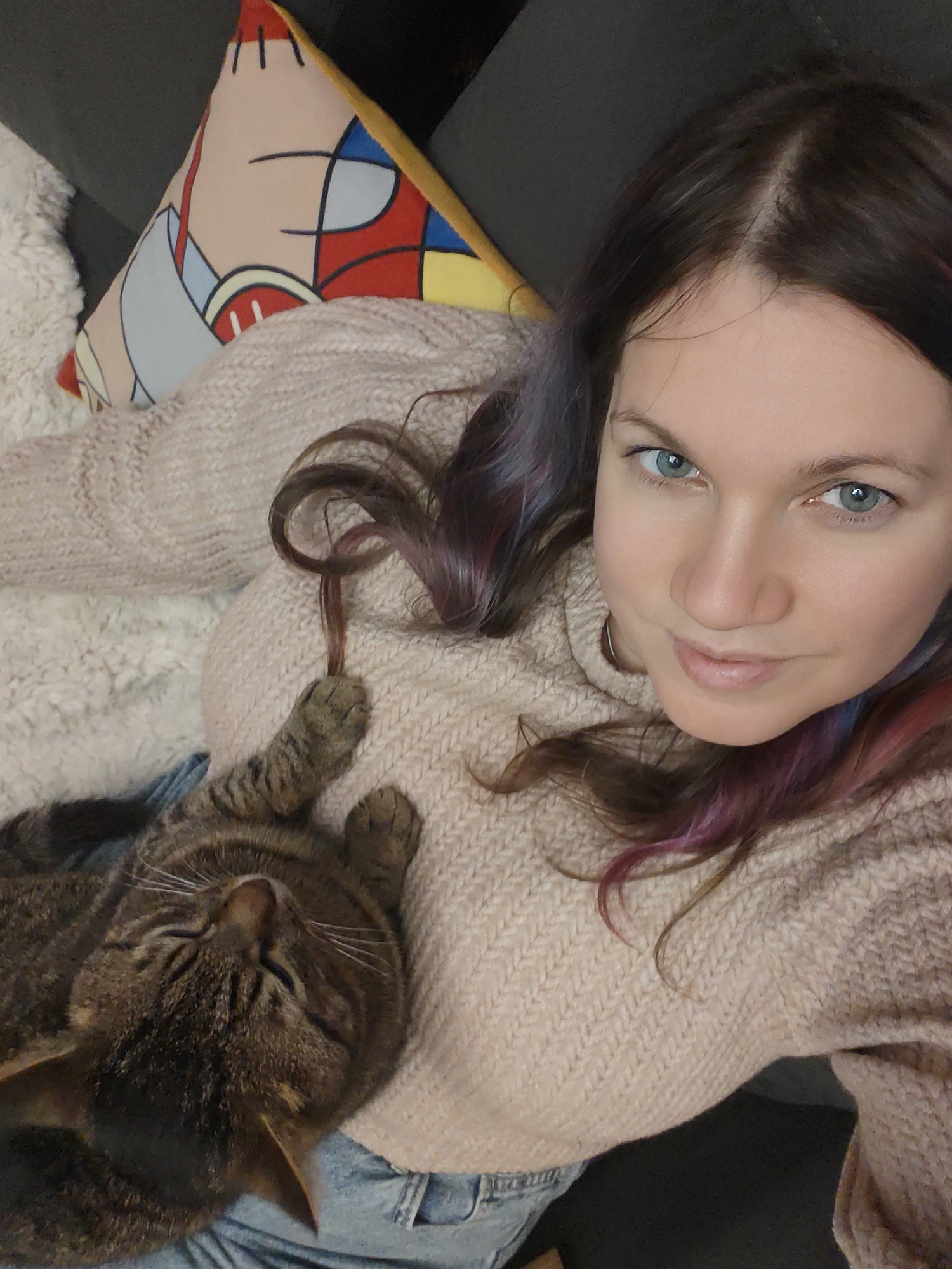 A woman is seen taking a high-angle selfie while lounging indoors with a tabby cat. She looks directly at the camera with blue-green eyes and a relaxed, slight smile. Her dark brown hair, with subtle purple or pink highlights, frames her face and shoulders. She wears a light beige or peach-colored chunky knit sweater, and light blue jeans are visible around her waist.
A brown tabby cat with dark stripes is lying contentedly on the woman's stomach and chest, seemingly asleep or very relaxed, with its eyes closed and head tilted back. Its front paws rest gently on the woman's sweater. White whiskers are visible on the cat's snout.
The setting suggests a comfortable indoor environment, likely a living room or bedroom. Behind the woman, a dark grey surface, possibly a sofa or headboard, is visible. A fluffy, cream-colored blanket or throw covers part of the surface and the woman. To the upper left, a decorative cushion with an abstract, geometric pattern in red, blue, yellow, and light beige colors adds a splash of brightness. The image conveys a peaceful and affectionate interaction between the woman and her pet. No visible text is present in the image. The lighting suggests it is daytime.