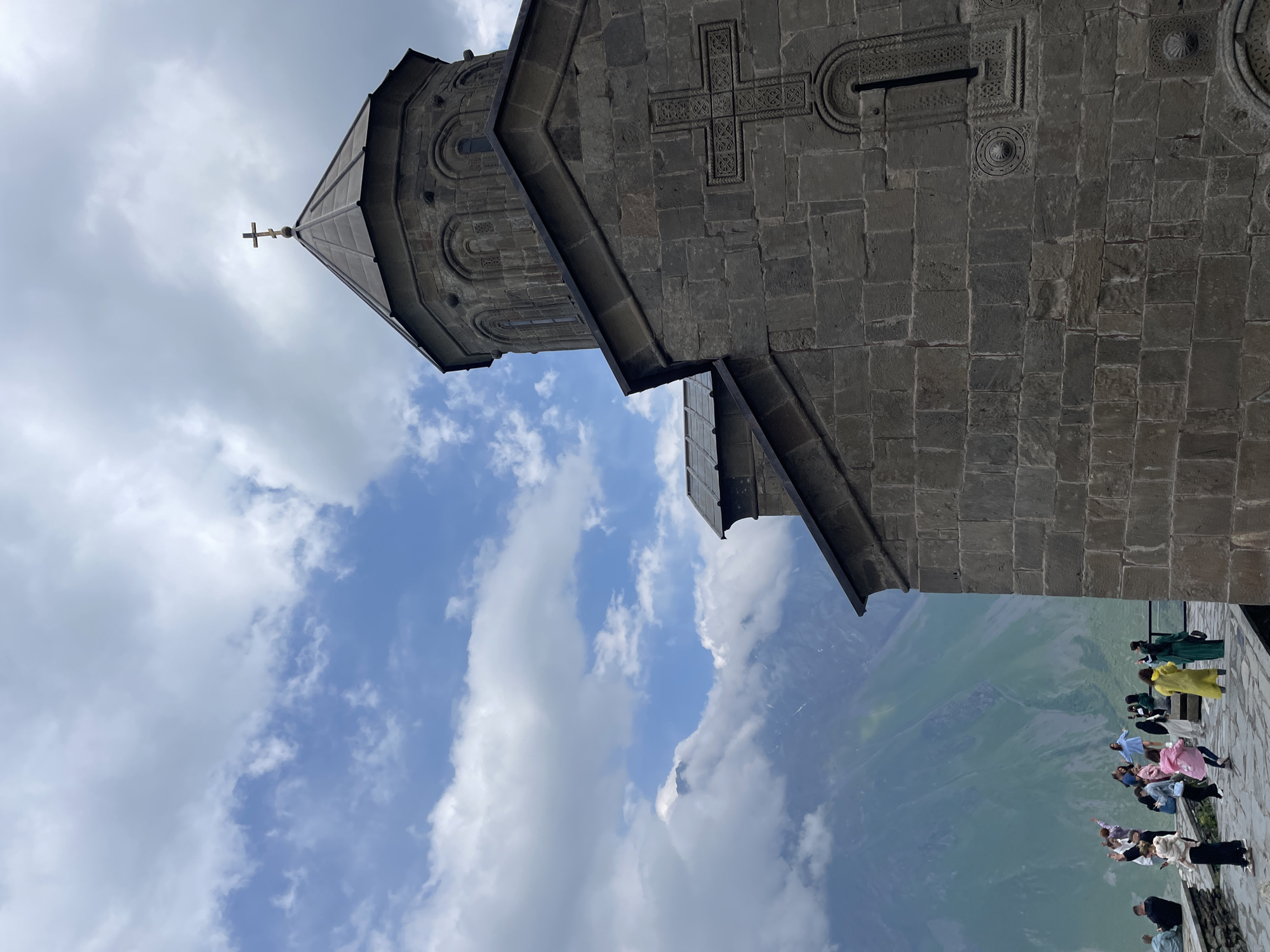 The image shows Gergeti Trinity Church, a 14th-century Orthodox church located in Stepantsminda, Georgia. The church is situated on a mountaintop overlooking the Greater Caucasus Mountains. The exterior of the church is made of rough-hewn stone blocks. A prominent feature of the church is a large, intricately carved cross on its facade. The dome of the church is conical and topped with a cross.
In the foreground, a group of tourists are gathered on a stone terrace. They are dressed in casual clothing and appear to be taking photographs and admiring the view. The background is dominated by a dramatic mountain landscape, with lush green slopes partially obscured by dramatic clouds. The sky above is a mix of blue and white clouds, suggesting it is a partly cloudy day. The overall scene depicts a historical landmark in a breathtaking natural setting, with visitors enjoying the scenery.
