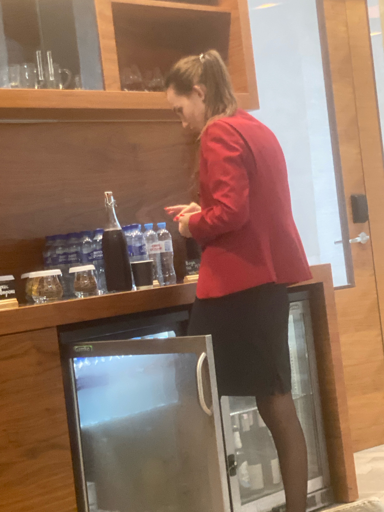 A woman in a red jacket and black skirt is standing in what appears to be a hotel lounge or bar area. She is reaching for something on a wooden shelf that holds various bottles of water, small jars, and what looks like a dark beverage in a stoppered glass bottle. To her left, there is a mini-fridge with its door open, revealing a shelf with some items. The environment suggests an indoor setting with wooden cabinetry and a glass-paneled door on the right. The image's quality is somewhat low, making finer details difficult to discern. The location is specified as Minsk, Belarus.