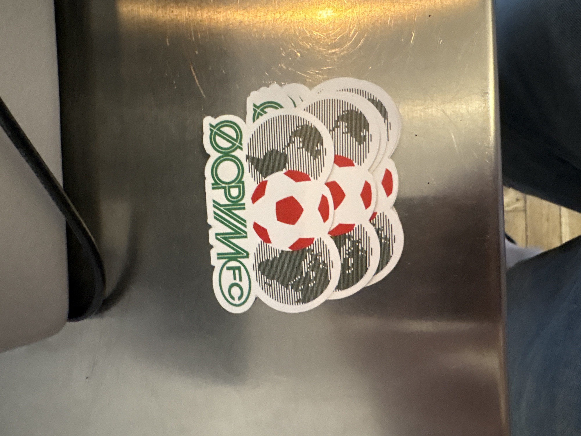 A sticker is affixed to a metallic surface, likely a refrigerator or locker, in a close-up shot. The sticker features a cluster of circular designs, each containing a stylized representation of the Earth with a soccer ball. The Earth is depicted with a dark grey silhouette, overlaid with thin black vertical lines, against a white background. Two prominent soccer balls, one in the foreground and another slightly behind it, are colored white with red pentagonal panels.
To the left of these circular designs, the text "ØOPM FC" is written vertically in bold green lettering with a white outline. The "Ø" character is a circled slash, and the "O" in "FC" is also enclosed in a green circle. The sticker is layered, with multiple circular elements stacked behind one another, creating a sense of depth.
The metallic surface shows some minor scratches and reflections, indicating it's a common household or public object. The background to the right is blurred and appears to be dark fabric, possibly clothing, and a hint of wooden flooring. The overall lighting suggests an indoor environment. No people are visible in the frame. The context of Belgrade, Serbia, is provided externally.