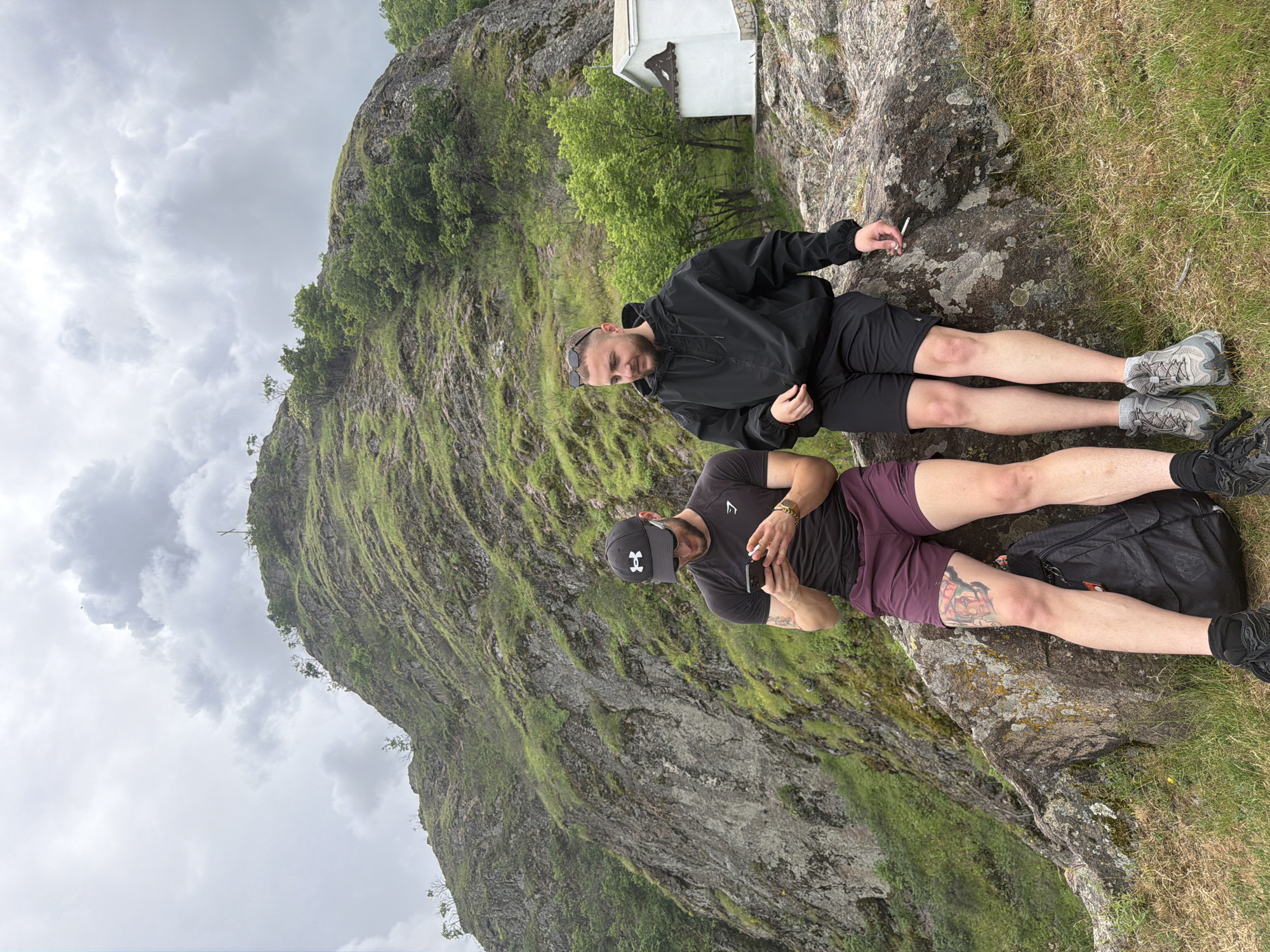 Two men are reclining on a rocky, grassy area in Ustina, Bulgaria, with a large rock formation behind them and a cloudy sky overhead. The first man, wearing a black jacket and shorts, is lying with his head near a small white building on the rock. He holds a cigarette in his left hand. The second man, wearing a black Under Armour hat, a black T-shirt, and maroon shorts, is holding a cigarette in his right hand and his phone in his left hand. He has a tattoo on his left thigh. A black backpack rests near his feet. The rock formation is covered in green vegetation.