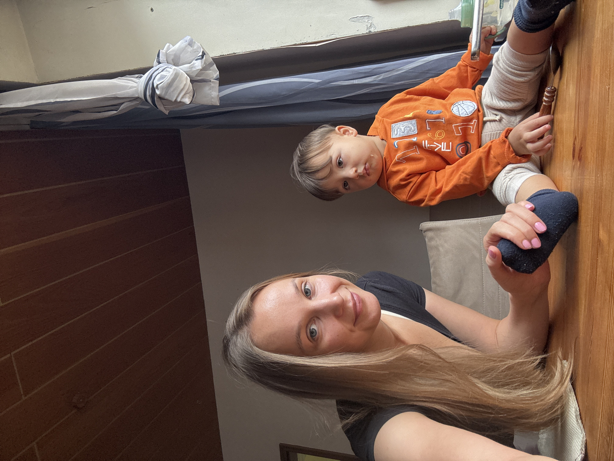 A medium shot captures a woman and a young child sitting at a wooden table in a room. The woman, with long blonde hair, is on the left, wearing a black t-shirt. She is leaning on her arm, holding a dark blue sock with her right hand, her nails painted pink. The child, on the right, is wearing an orange hooded sweatshirt with numbers and a basketball graphic, and light-colored pants. He is looking away from the camera with a slightly drooling mouth. Visible objects on the table include a spoon and a wooden object. The background features a grey wall and a window with blue and white striped curtains tied back. The ceiling is made of dark wood planks. The scene is consistent with an indoor setting in a residential property in Überherrn, Germany.