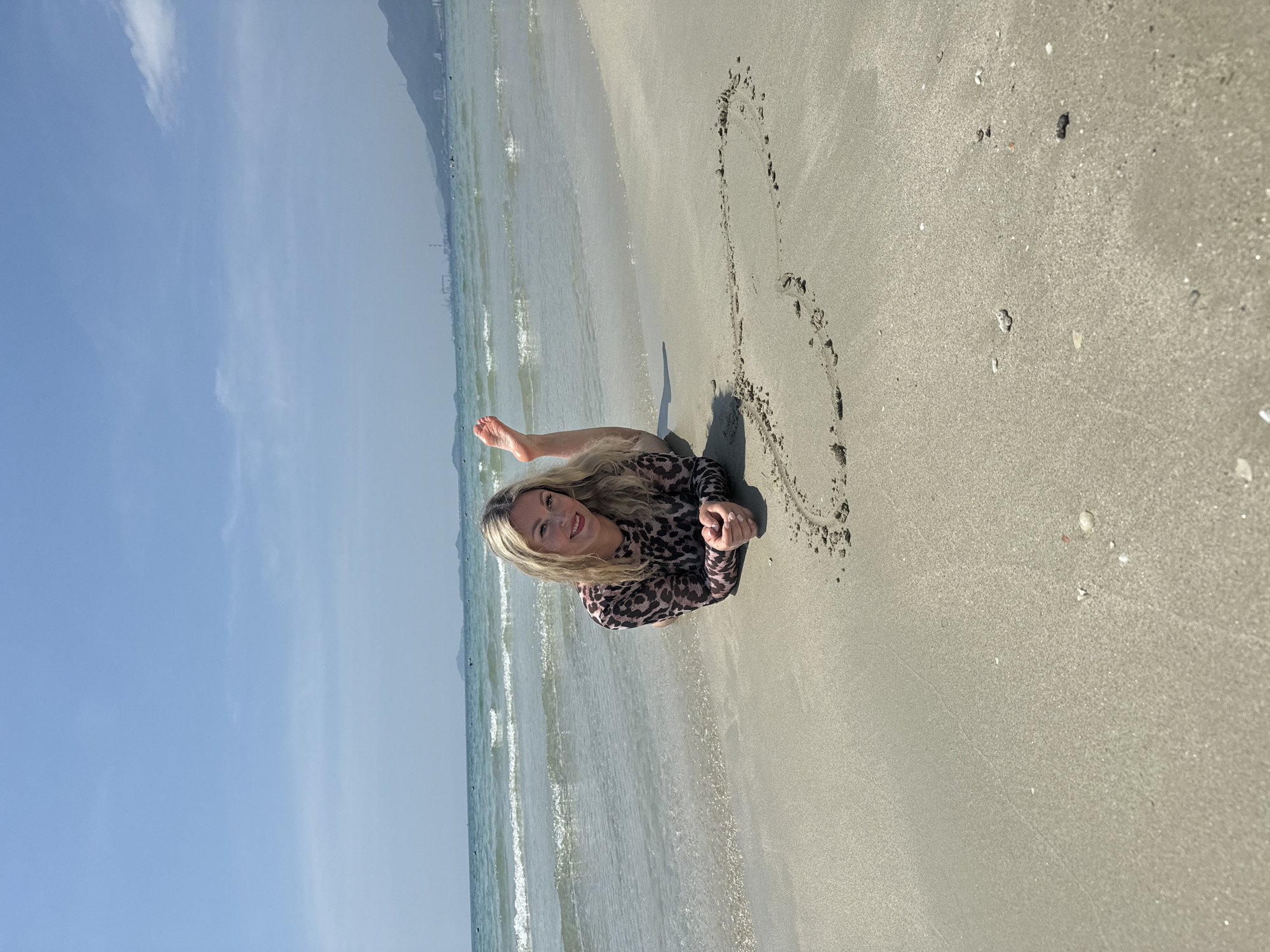 The image captures a woman lying on a sandy beach in Dong Ninh Hoa Ward, Vietnam. She is positioned near the water's edge, with gentle waves washing onto the shore. The woman is wearing a leopard-print long-sleeved shirt and has her hair down. Next to her, a heart shape is drawn in the sand using footprints. The sky above is a clear blue, indicating a sunny day. In the background, there are hints of buildings and a mountain range. The sand is dotted with seashells and debris.