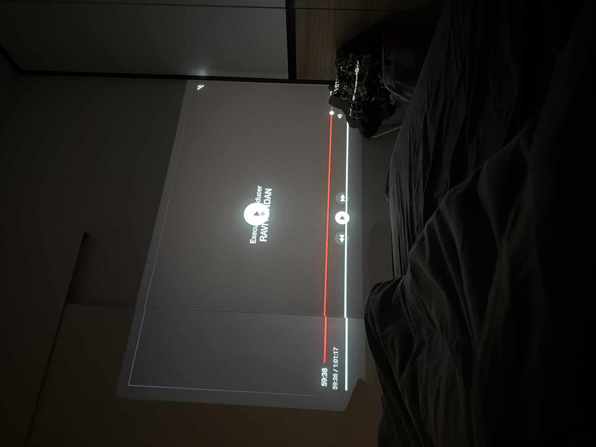 Here is a description of the image. The image shows a television screen displaying a YouTube video paused at 59:38 of a 1:01:17-long video. The video credits list Ravi Nondan as an executive producer. The screen is dark, with a red progress bar indicating how far into the video it is. The furniture and bedding are draped with dark fabric. The scene appears to be indoors, possibly in a bedroom, given the presence of a bed with its bedding. The location in the city is unknown.