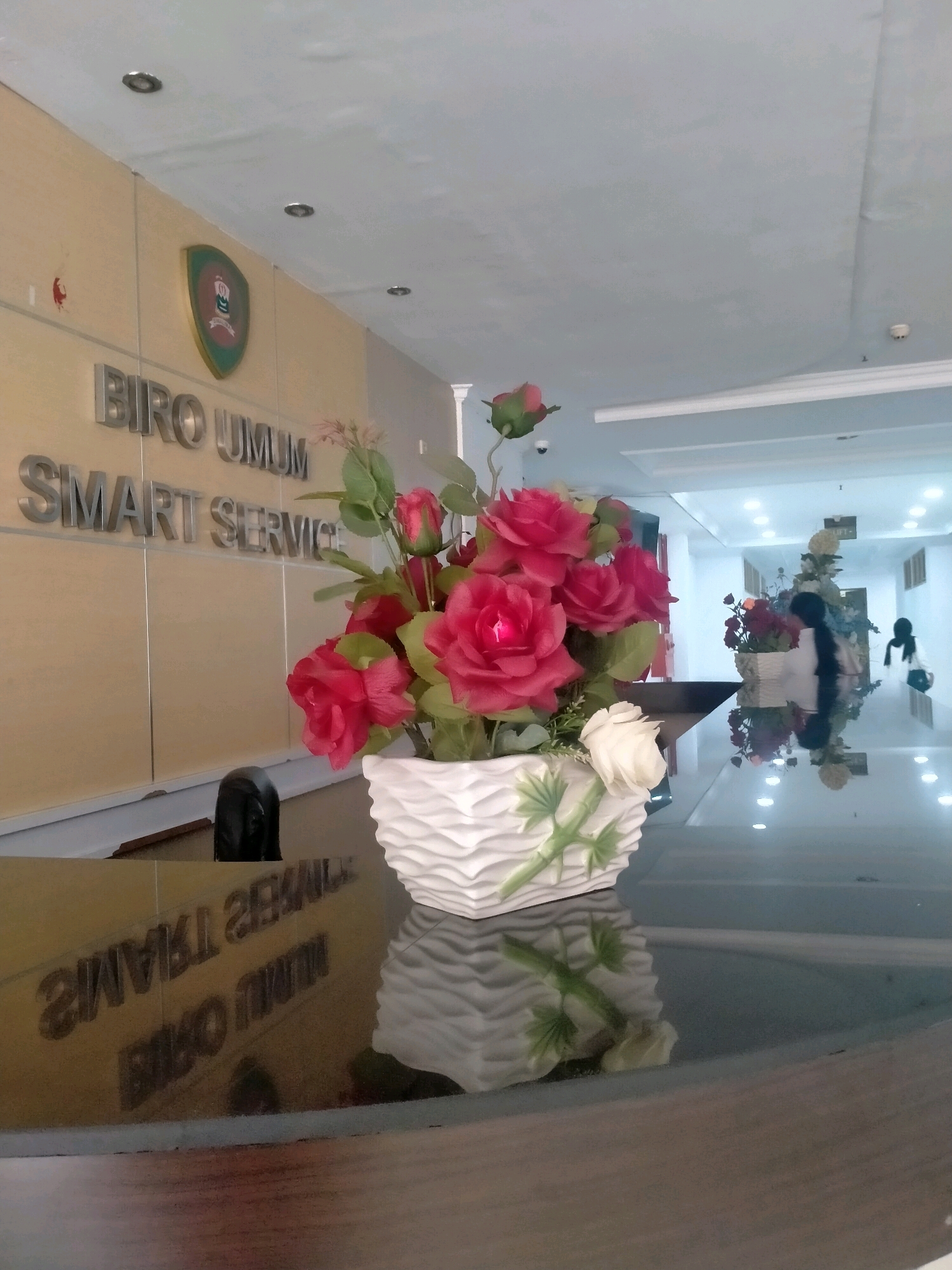 The image captures a close-up view of a floral arrangement on a reflective surface, likely a table or counter, in what appears to be an office lobby. The artificial flowers, predominantly in shades of red and pink with some white accents, are displayed in a white, textured vase. In the background, metallic lettering on a beige wall reads "BIRO UMUM SMART SERVICE," indicating a government or administrative office. A shield-shaped logo with green and red elements is positioned above the text. The environment is brightly lit with ceiling lights. The reflective surface shows the mirrored image of the flowers and the text, as well as blurred figures of people in the distance, one wearing a black headscarf. The scene is consistent with the interior of an office reception area in Ambon, Indonesia, given the presence of the Indonesian language on the signage.