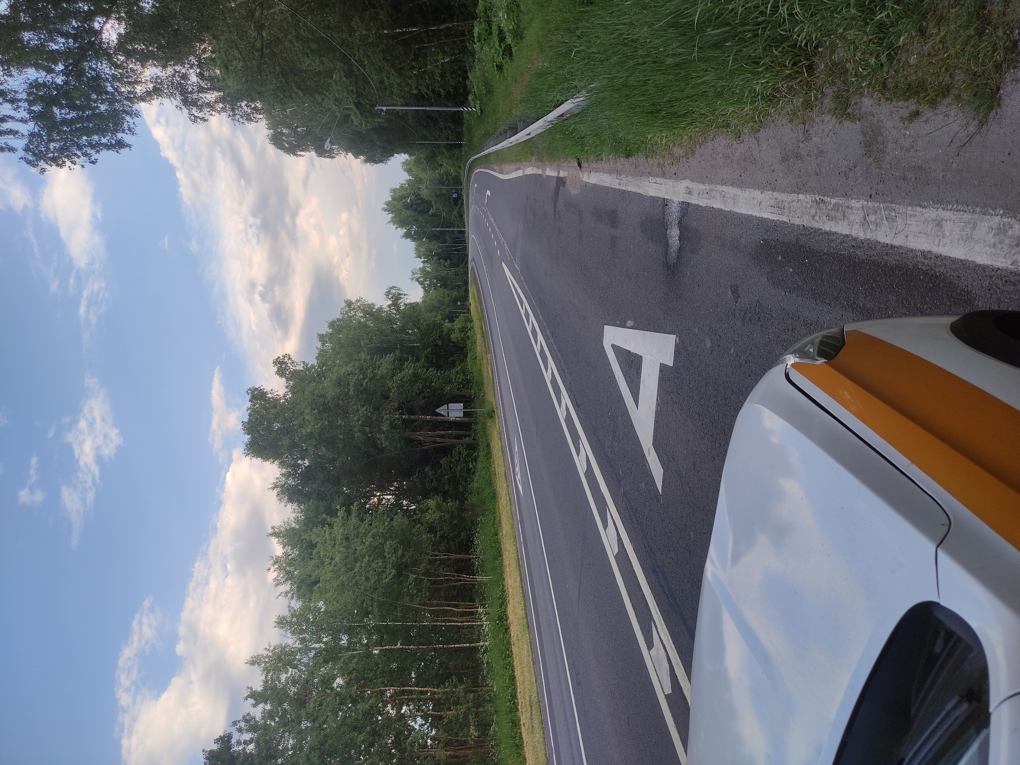 The image shows a road in a wooded area, with the hood of a white car with orange stripes in the foreground. The road has lane markings and traffic signs. The sky is cloudy, with some patches of blue. The scene is calm and peaceful. It is daytime. The location is a road in Kopylovo, Russia.