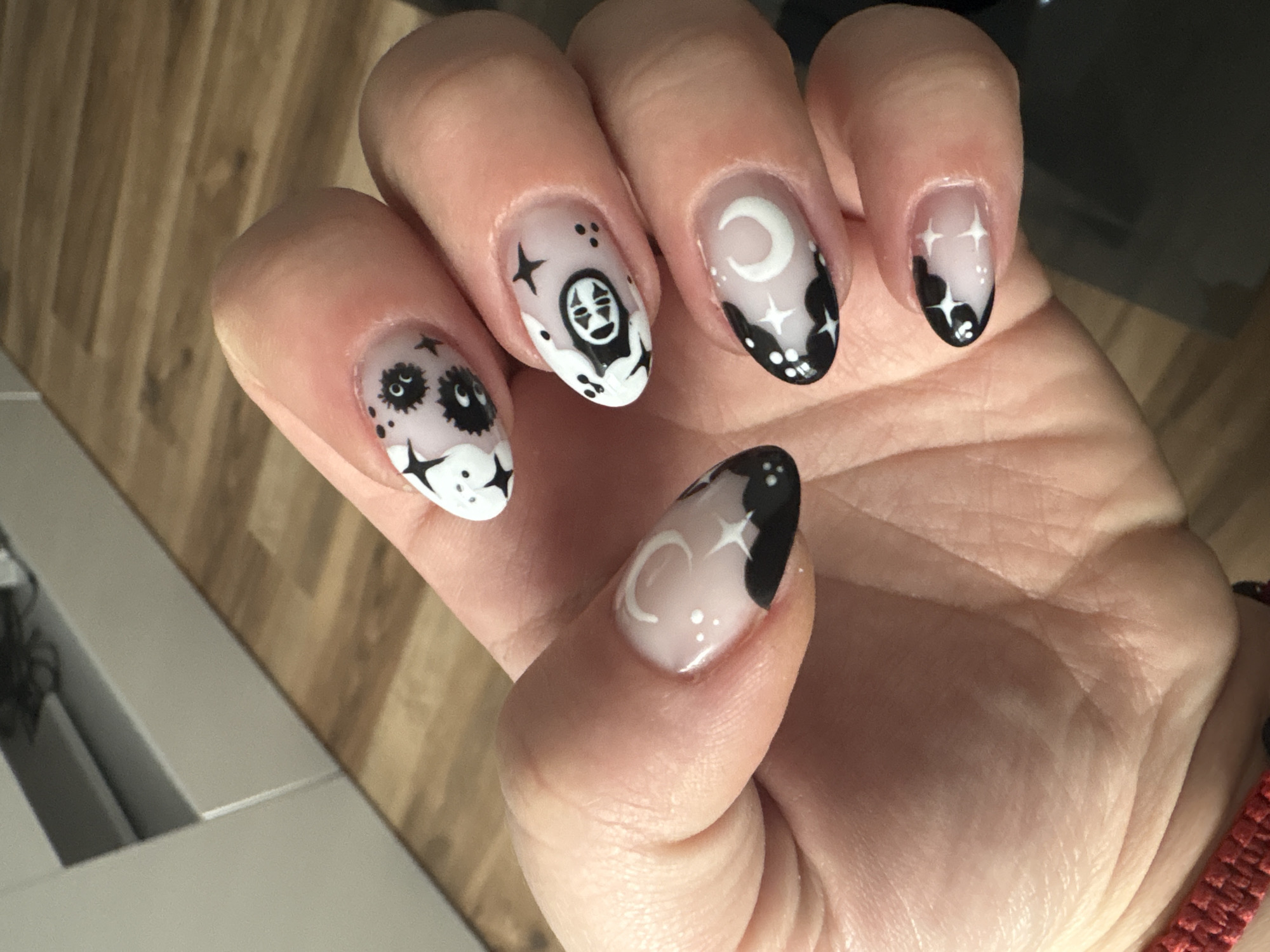 A person's hand is displayed with distinctive nail art on each finger. The nails appear to have a clear base coat with black and white designs. The thumb features a crescent moon with clouds and stars, the index finger features the character "No Face" from the film Spirited Away, the middle finger has a crescent moon with clouds and stars, the ring finger shows stars and clouds, and the pinky finger shows stars and clouds. The background shows the wooden floor and some furniture. There is no explicit information or indication in the image connecting it to the city of Plovdiv, Bulgaria.