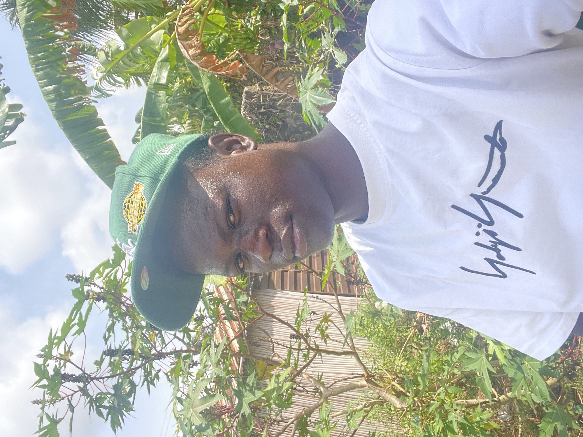 A photograph captures a person in an outdoor setting, surrounded by lush greenery suggestive of a tropical climate. The individual, with dark skin and a slightly serious expression, is wearing a green baseball cap with a gold emblem and a white t-shirt with a black design. The background includes various plants with broad leaves and what appears to be a corrugated metal structure partially visible. The location is implied to be Badagry, Nigeria, based on the prompt. The lighting suggests a bright, sunny day.