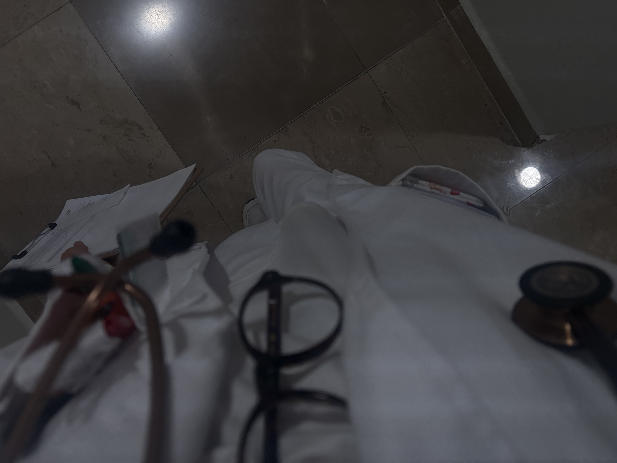 The image captures a first-person, low-angle perspective looking down at a scene involving a person in a medical setting. The primary subject is a person wearing a white lab coat or scrub top, partially visible from the waist down, with their legs and torso covered by the white fabric.
Resting on the white attire are several medical instruments. A stethoscope, featuring copper-colored tubing and black ear tips, is prominent in the foreground on the left, extending towards the upper left. Another part of a stethoscope, likely the chest piece, is visible on the right side. A pair of black-framed glasses is placed near the center, slightly to the left, on the white fabric. On the far left, a stack of white papers or a notebook is partially visible.
The background consists of a dark, tiled floor, possibly made of marble or ceramic, with visible grout lines. The floor reflects several light sources; a large, bright, circular reflection is seen in the upper left, and a smaller one appears towards the lower right. The overall lighting is dim and diffused, suggesting a nighttime setting or a dimly lit indoor environment.
The scene conveys the atmosphere of a medical professional's immediate surroundings, possibly during a break or after a shift, with essential tools laid out. No specific activity is actively taking place; it's a static snapshot. There is no visible text in the image. Given the provided context, this scene is located in Puebla, Mexico.
