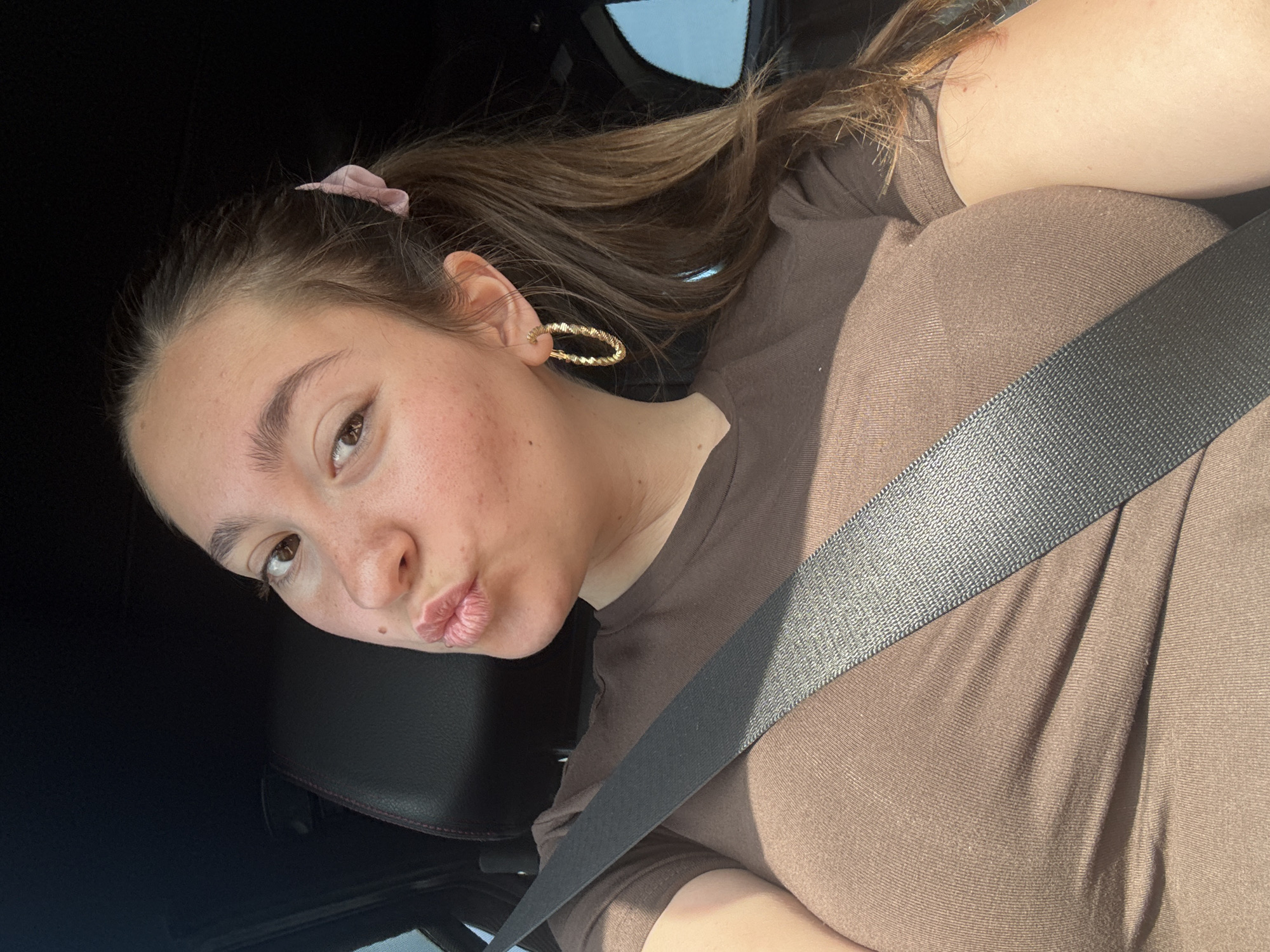 A young woman is taking a selfie in a car. She is wearing a brown t-shirt and a seatbelt. Her hair is brown and pulled back in a ponytail. She has hoop earrings and a pink scrunchie in her hair. She is puckering her lips. The background is dark, but appears to be inside a car. The image is taken in Polsko Padarevo, Bulgaria.