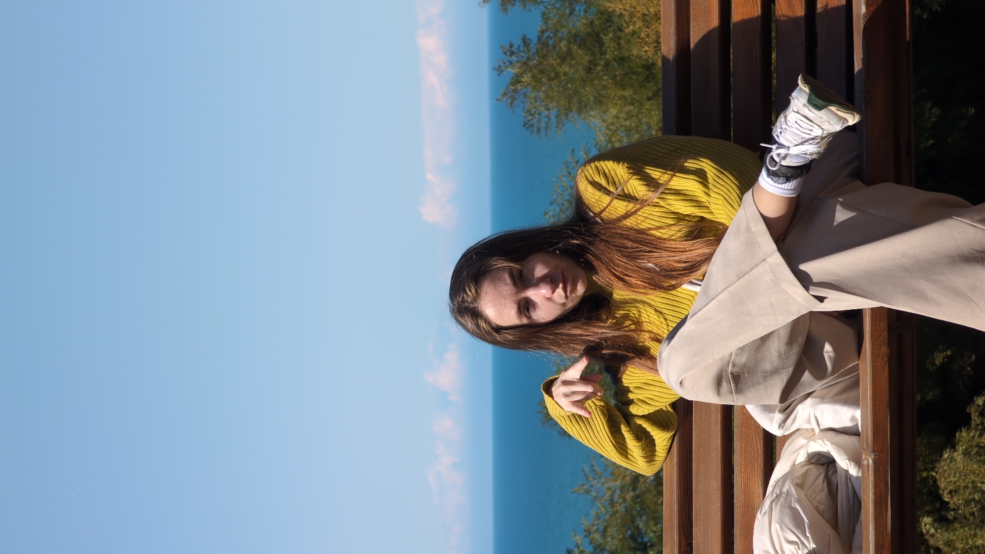 A woman is sitting on a wooden bench in Batumi, Georgia. She is wearing a mustard-colored sweater, beige pants, and white sneakers with black socks. She is facing the camera, with her legs stretched out in front of her. The bench overlooks the Black Sea, with a clear blue sky in the background. There are trees and vegetation on either side of the bench. A jacket is draped over the bench near the woman's legs.