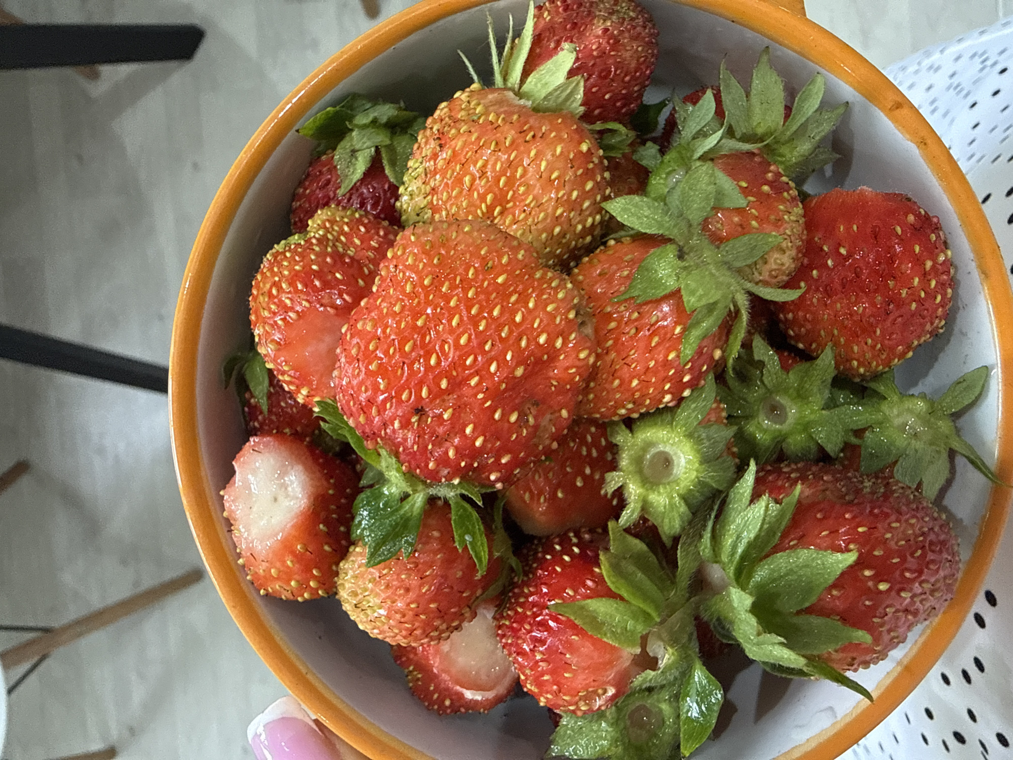 The image shows a close-up of a bowl filled with fresh strawberries. The bowl is orange on the outside and white on the inside. Some strawberries are whole, while others are cut in half, revealing their white interior and small seeds. The strawberries are ripe and red, with green leafy tops. The background is blurred, showing parts of furniture and a floor, suggesting an indoor setting. There is no discernible information about the location in the city of Октябрьское, Russia, as the image is cropped and focuses solely on the strawberries in the bowl.