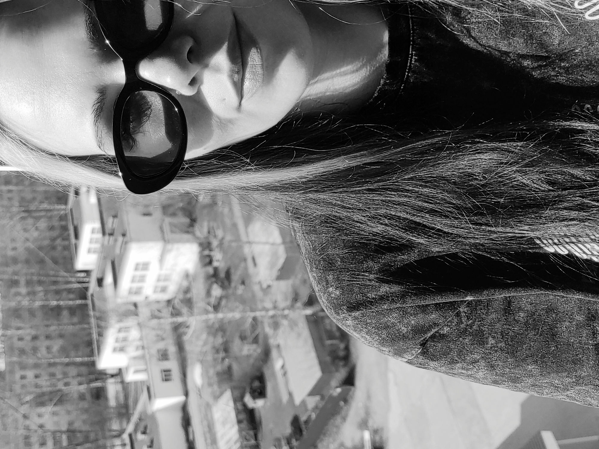 This black and white, close-up portrait features a person, likely female, with long, light-colored hair, wearing dark, oval-framed sunglasses. The subject's face is shown from the forehead to the chin, tilted slightly upwards and to their right. The sunglasses obscure the eyes but show reflections of the bright environment. Her lips are slightly parted, revealing a subtle texture, and a defined shadow falls across her upper lip. Strong highlights on her forehead, cheek, and hair, contrasted with deep shadows on her neck and under the sunglasses, indicate bright, direct sunlight, suggesting a clear, sunny day. Her hair, appearing long and flowing, extends across her shoulder and the left side of the frame. She is wearing a textured garment, possibly a denim jacket or shirt, on her right shoulder and upper arm. The background is softly blurred, revealing an urban or suburban landscape with several buildings and distant architectural structures, suggesting the photo was taken outdoors from an elevated perspective. The overall mood is casual and relaxed. No visible text is present in the image.