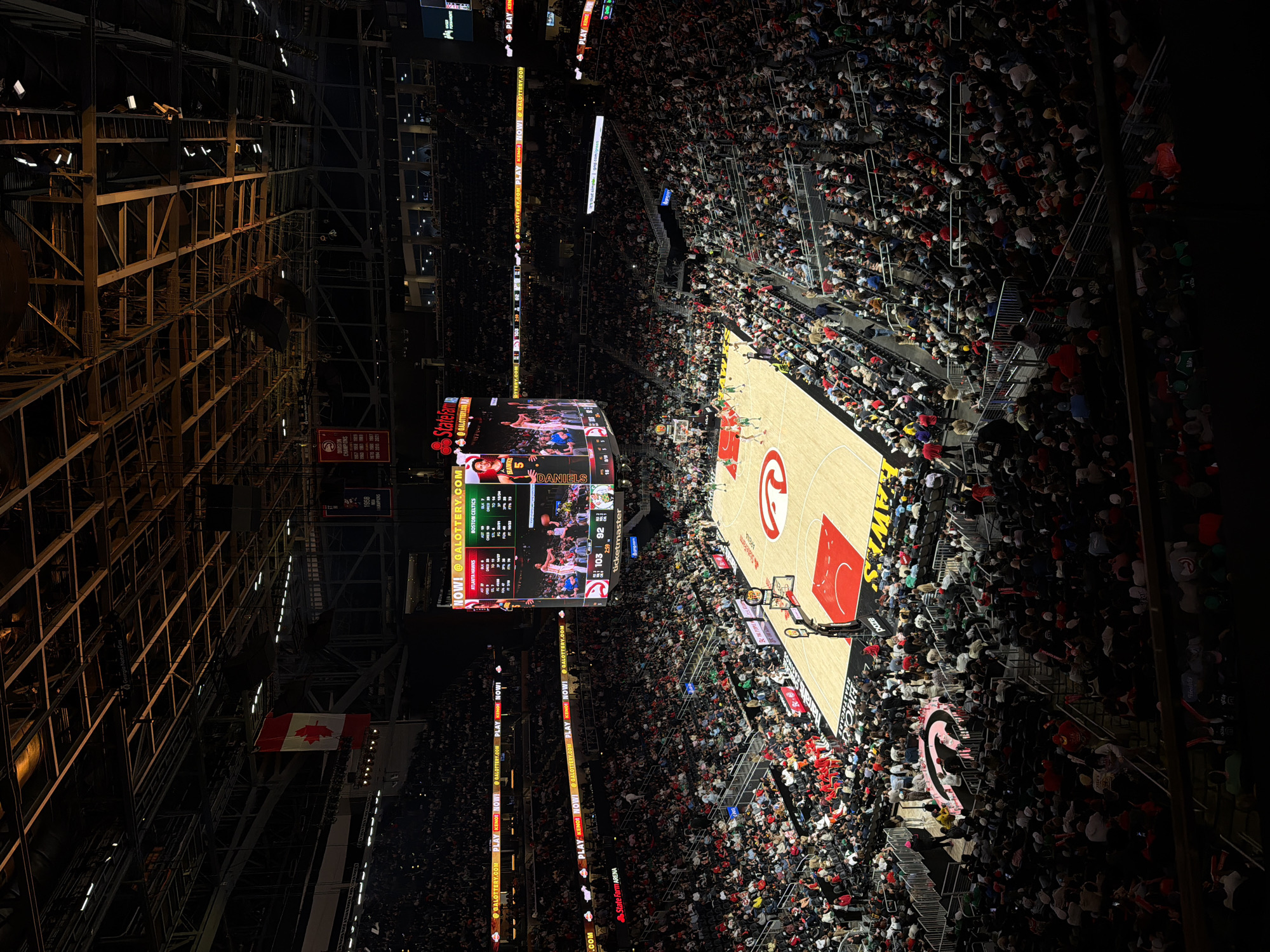The image depicts a high-angle view inside the State Farm Arena in Atlanta, United States of America, during a basketball game between the Atlanta Hawks and the Boston Celtics. The basketball court is visible, with players in action. Surrounding the court, the stands are filled with spectators. A large video scoreboard hangs above the court displaying the game score and player statistics. Structural elements of the arena's ceiling and lighting system are also visible, as is a Canadian flag.