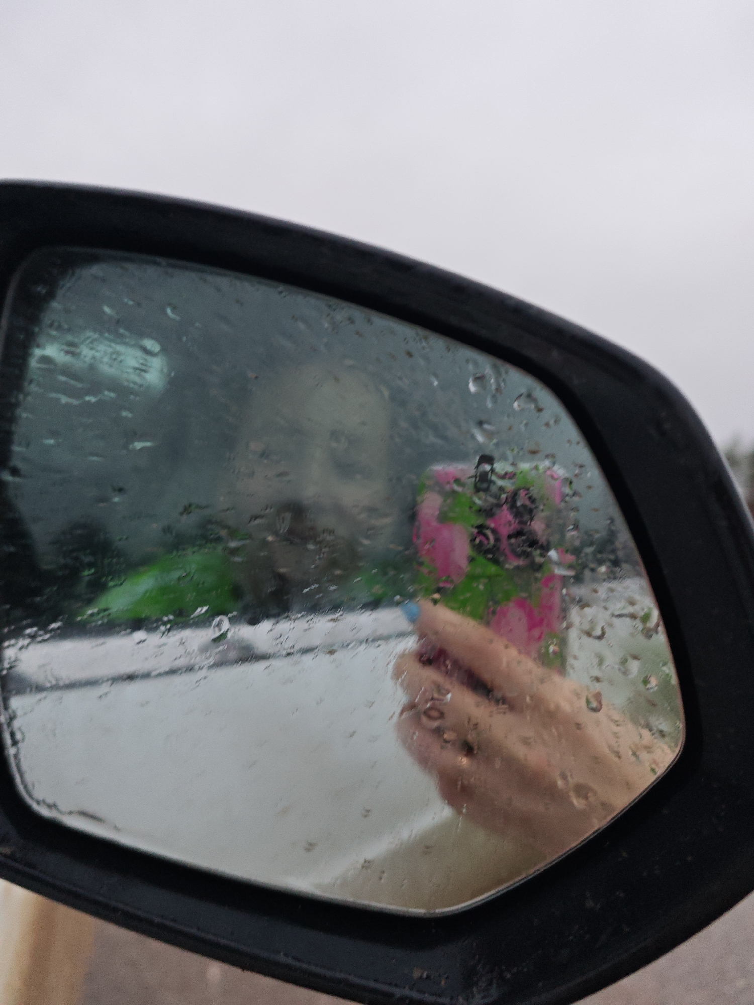 The image depicts a side view mirror of a car. Raindrops are visible on the mirror's surface, obscuring the reflection. Within the reflection, a person is holding a pink and green item, possibly a bouquet or a decorated object. The person's face is partially visible in the background of the mirror's reflection, although it is somewhat blurred. The overall scene appears to be taken in overcast or rainy weather. No specific landmarks or details confirm the location as Dubovaya, Russia.