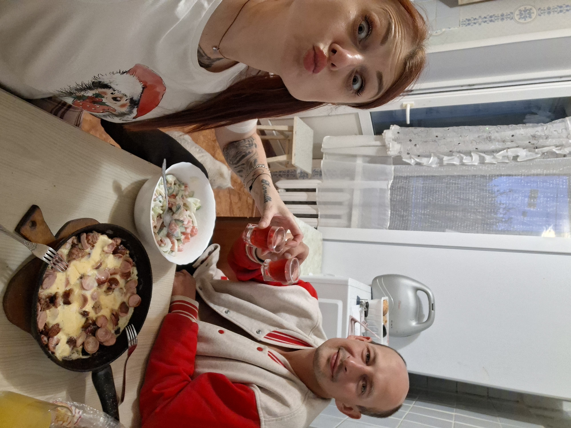 The image depicts an indoor scene, likely a kitchen or dining area, with two individuals seated at a table. A woman in the foreground, with reddish-brown hair, is making a kissy face towards the camera. She is wearing a white t-shirt with a graphic of a hedgehog in a Santa hat holding a strawberry. A man, to her right, is smiling and holding up two small shot glasses filled with a red liquid. He is wearing a varsity-style jacket with a cream-colored body and red sleeves. On the table in front of them, there is a bowl of salad and a frying pan containing food, possibly eggs and sausages, with two forks. The environment suggests a casual meal or celebration. The location is identified as Забалоцкі сельскі Савет, Belarus.