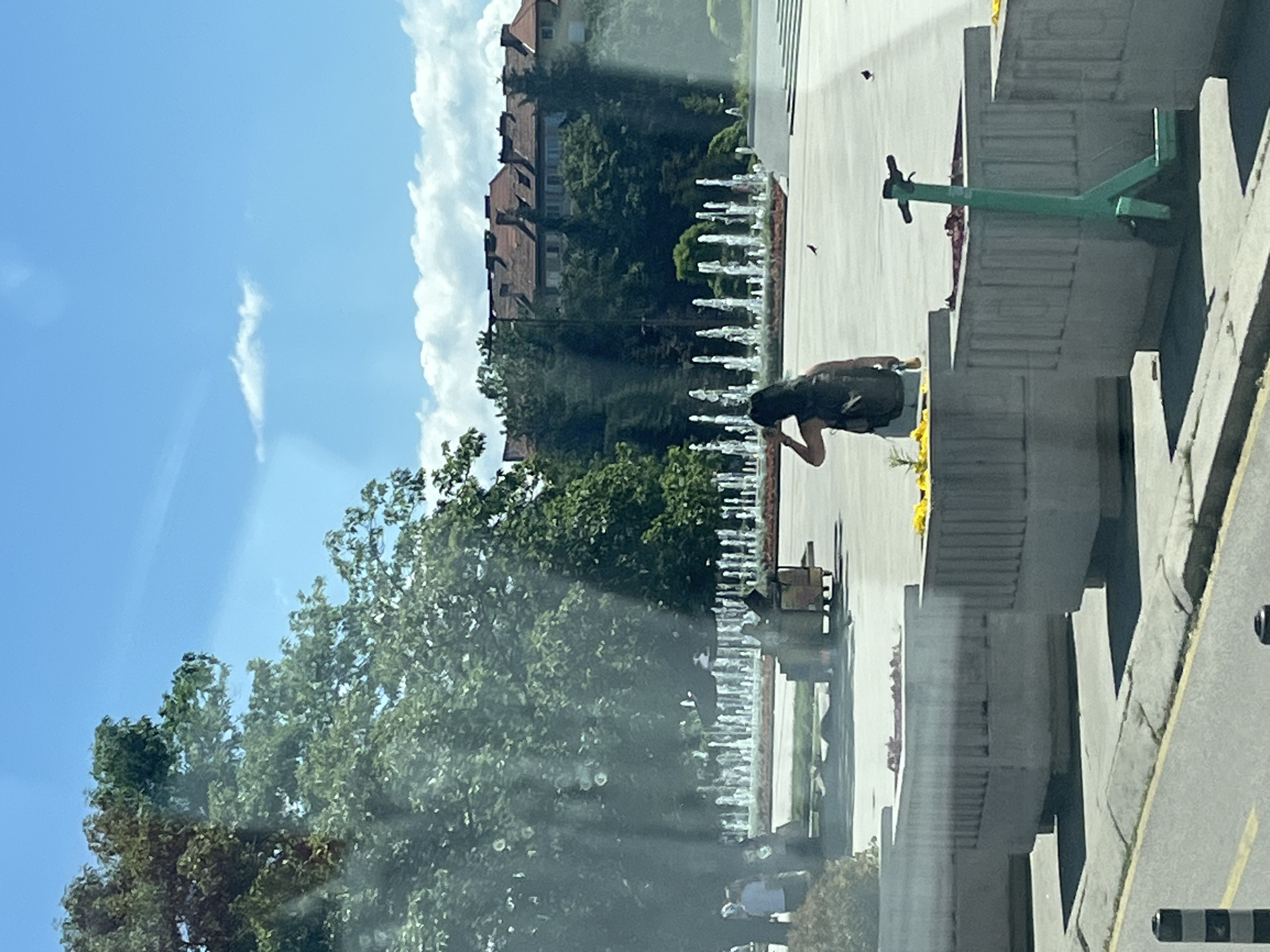 An outdoor scene shows a woman with a backpack standing with her arm raised to her head, facing away from the camera. She is in a plaza with fountains in the background. In the foreground, there are large concrete planters, one of which has a green electric scooter leaning against it. Several trees are visible on the left and in the middle ground, and buildings with red roofs are in the background. The sky is blue with scattered clouds.