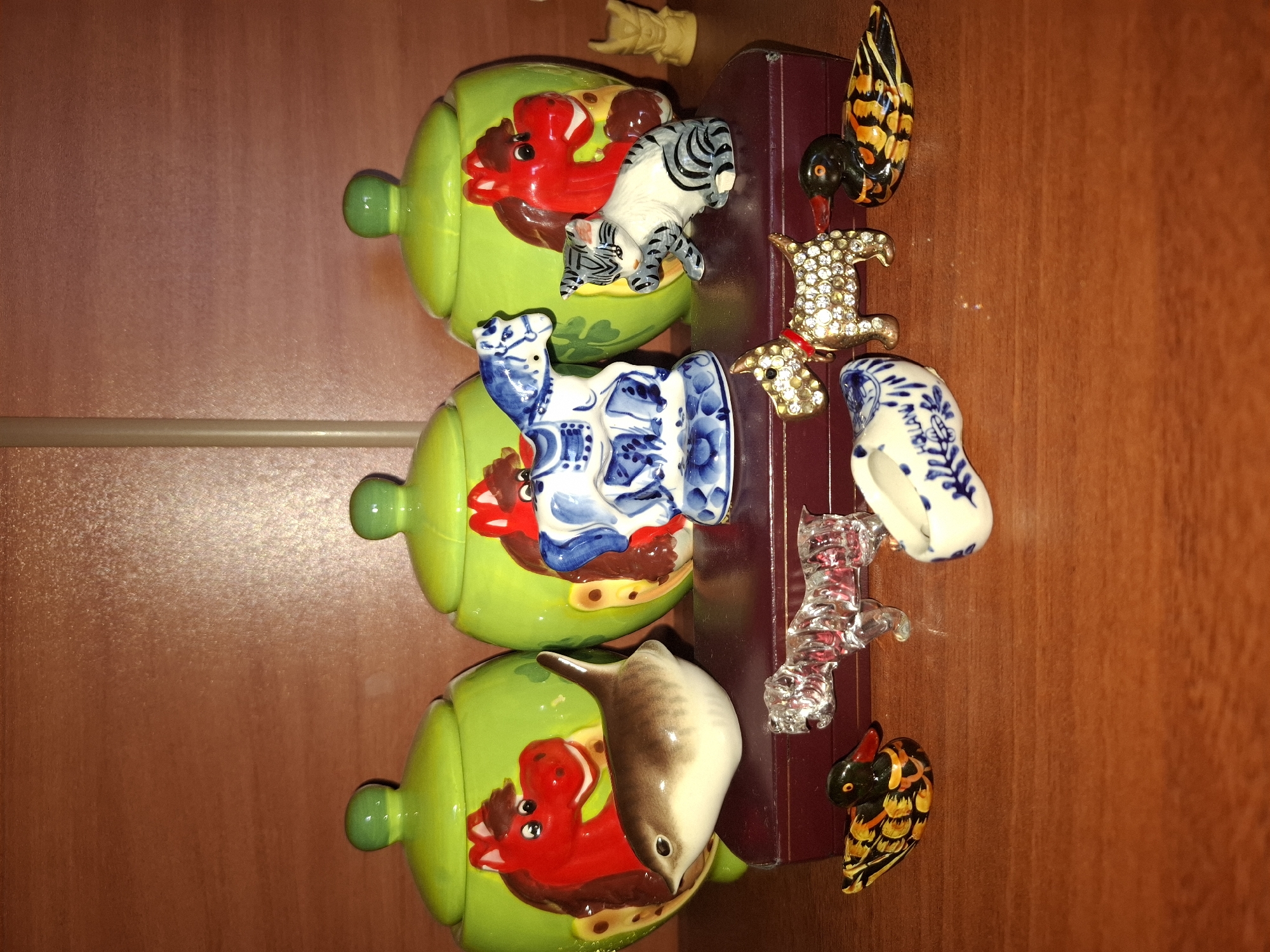 A close-up shot features a collection of decorative ceramic and glass figurines arranged on a wooden surface. In the background, three lime-green ceramic jars with rounded lids are visible. Each jar is adorned with a painted red horse head on its front.
In the middle ground, positioned on a dark red, rectangular box, are several figurines. To the left is a small ceramic bird with brown and white coloration. Next to it is a blue and white porcelain horse, followed by a white and gray ceramic cat.
In the foreground, on the wooden surface, are more figurines. To the left is a small duck figurine with black, yellow, and orange patterns. Beside it is a clear glass figurine of a dog. To the right of the glass dog is a small, white ceramic clog with blue floral patterns. Further to the right is another collectible dog figurine, this one appears to be made of metal and is encrusted with small clear stones, wearing a red collar. To the far right is another duck figurine, similar in pattern to the one on the left. A small, light yellow ceramic figure, possibly a mythological creature, is visible on the far right, behind the green jars.
The lighting in the image is somewhat dim, casting shadows and creating a slightly warm tone on the wooden surface. The background appears to be a tiled wall. The location is not specified as Almaty, Kazakhstan.