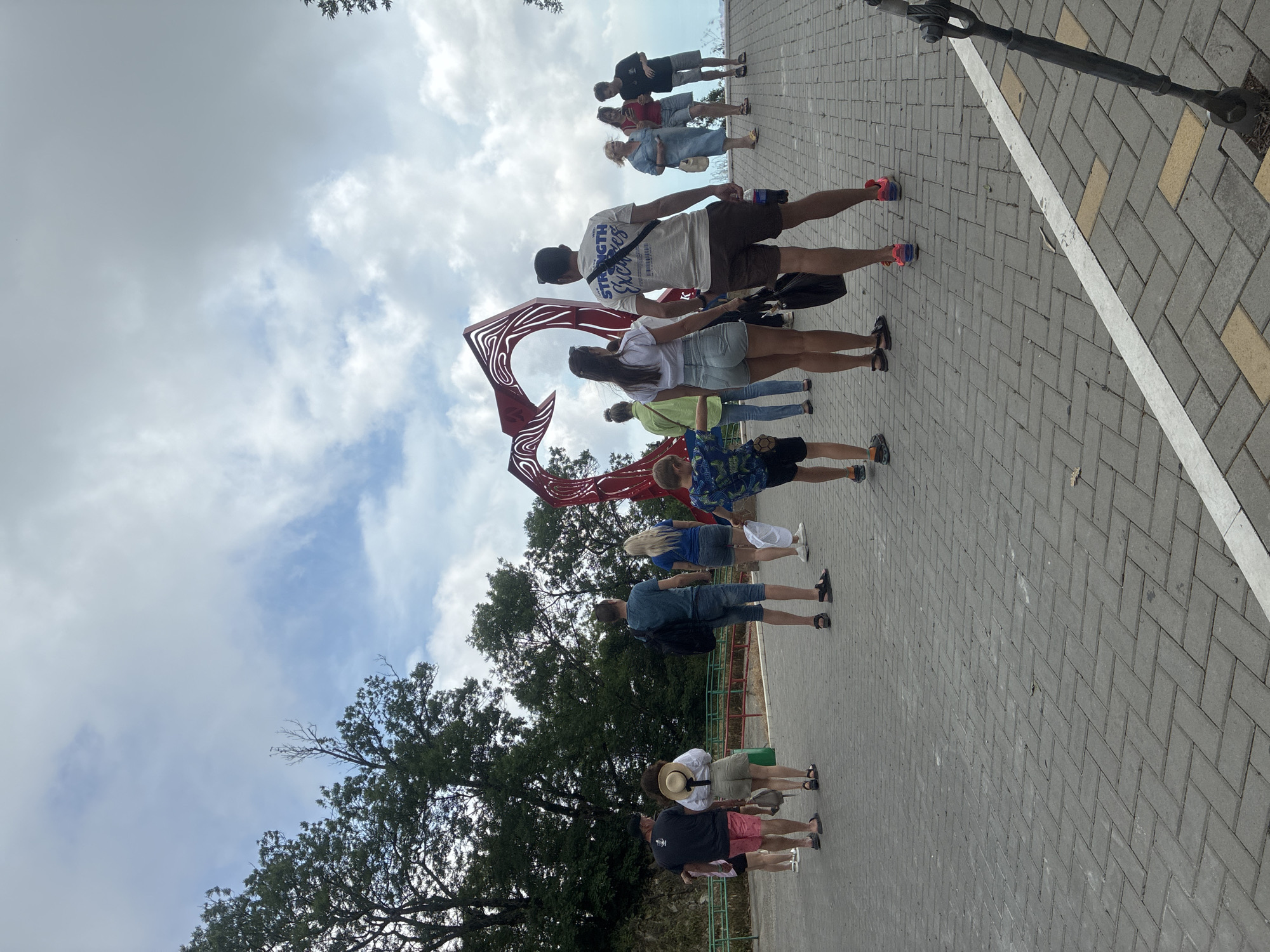 The image depicts an outdoor scene with a group of people gathered near a large, red, heart-shaped sculpture on a paved area. The sculpture is abstract and appears to be made of metal with swirling patterns. The people are casually dressed, suggesting a warm day. Some are looking at the sculpture, while others are walking or standing.
The location appears to be a tourist spot, likely a promenade or a viewpoint, given the sculpture and the presence of multiple individuals enjoying the scenery. The background features lush green trees and a cloudy sky.
This scene is consistent with the area around the "Heart of the World" sculpture located on the Gelendzhik embankment in Gelendzhik, Russia. This sculpture is a popular attraction, often visited by tourists and locals.
