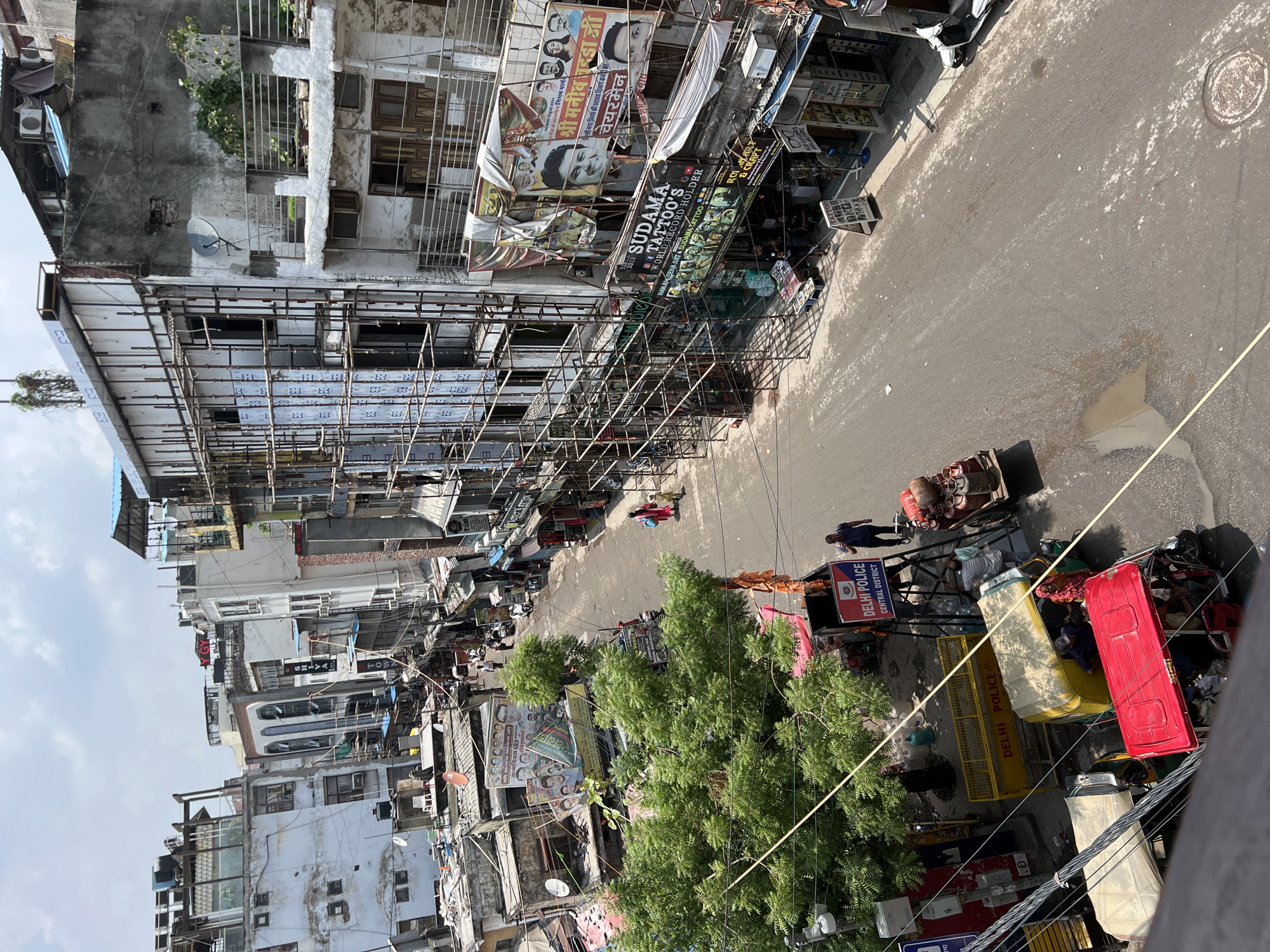 This is an aerial view of a busy street in Delhi, India, likely a commercial or mixed-use area. The scene depicts a narrow street lined with multi-story buildings, many of which appear to be undergoing construction or renovation, as evidenced by the scaffolding covering several facades. The buildings are a mix of older, somewhat dilapidated structures and newer constructions. Shops are visible on the ground floor of many buildings, with signs for businesses like "Sudama Tattoo's" and "Rohini Art & Craft."
The street itself is paved and shows signs of wear, with some puddles and debris. Several vehicles are present, including auto-rickshaws (one red and one yellow), a cart loaded with gas cylinders, and a scooter parked on the side of the road. Pedestrians are also visible, including a person in a blue shirt walking and two individuals, one in red and one in yellow, walking together on the sidewalk. A "Delhi Police Central District" sign is prominently displayed on a barricade.
The overall atmosphere suggests a bustling urban environment with a lot of activity. The weather appears to be clear and sunny. The architecture and street scene are characteristic of many older, densely populated areas within Delhi.