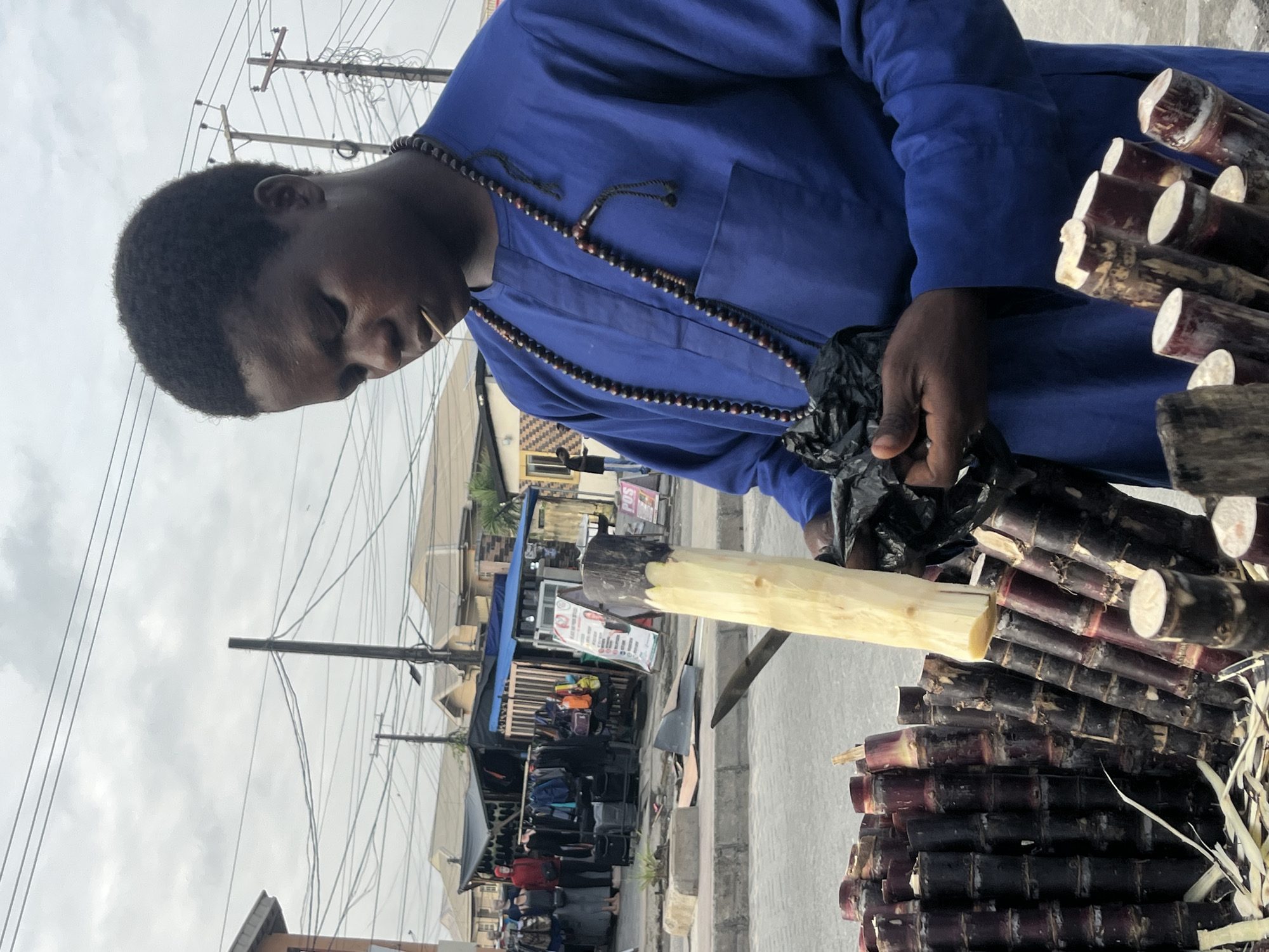 A dark-skinned person, likely male, wearing a blue dashiki and a beaded necklace, stands outdoors in what appears to be a marketplace in Ado, Nigeria. The person is holding a peeled section of sugarcane in their right hand and is looking down at it, with a toothpick in their mouth. In front of them, to the left, is a large pile of unpeeled sugarcane stalks. Behind the person, a market stall with various clothing items is visible, along with overhead power lines and buildings. The sky is overcast.