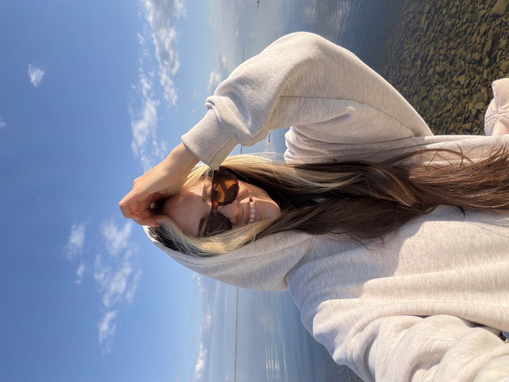 A young woman, wearing a light gray hooded sweatshirt and brown sunglasses, stands by a body of water in Kamyshin, Russia. Her hair is brown with blonde streaks. She is smiling, and her right hand is raised near her forehead. The water appears to be shallow with visible rocks. The sky is blue with scattered clouds.