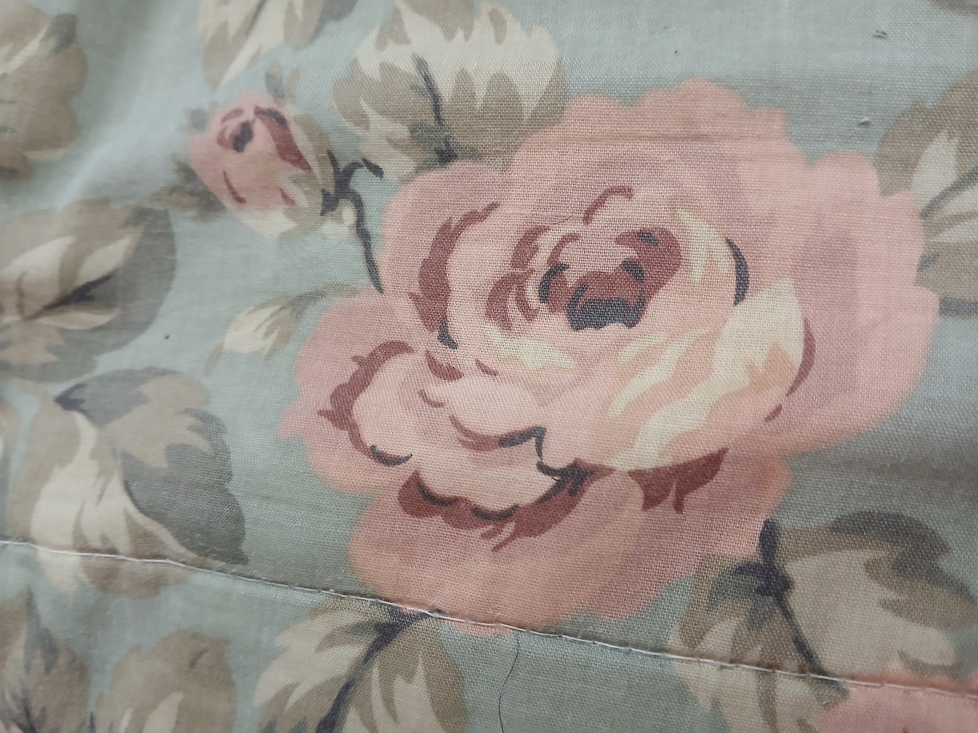 The image displays a close-up view of a floral fabric pattern. The pattern features large, muted pink roses with brown and cream-colored accents, interspersed with greyish-green leaves. A visible white stitch runs vertically through the center-left of the frame, indicating a seam in the fabric. The overall texture of the fabric appears to be woven cotton or a similar material, with a slightly matte finish. The lighting is soft and diffused, suggesting an indoor setting or an overcast day. No specific location within the city of Columbia, United States of America, can be determined from the visual information provided.