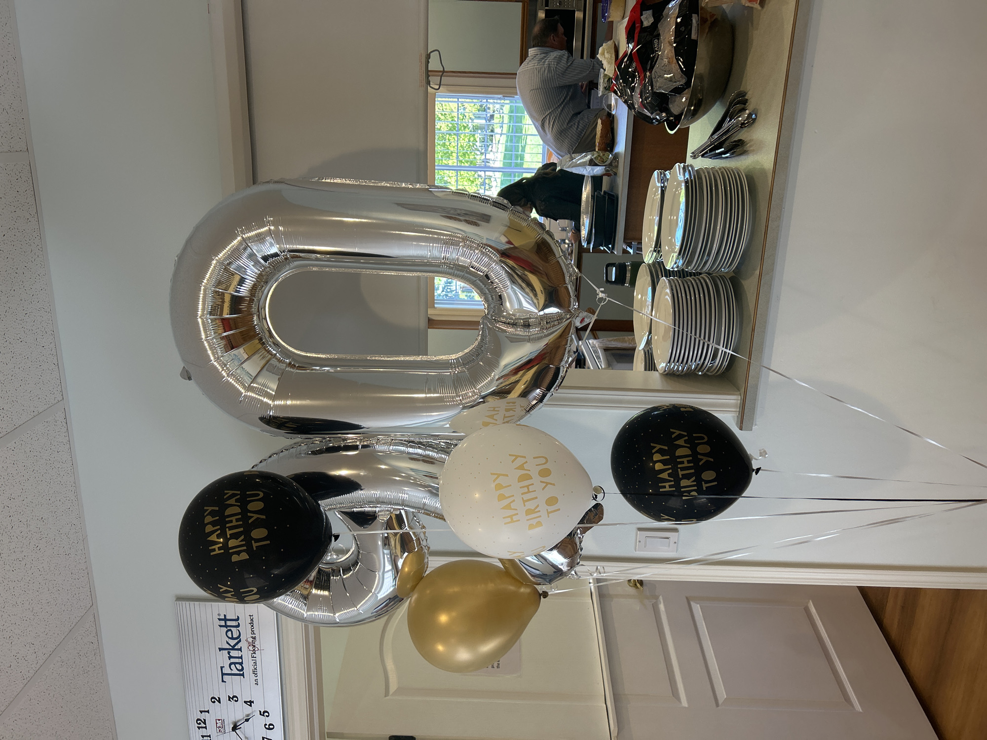 The image shows a cluster of balloons, including two large silver foil balloons shaped like the number "80", along with three smaller latex balloons. Two of the latex balloons are black with gold lettering that reads "Happy Birthday to You", and one is white with the same gold lettering. There is also a gold latex balloon. The balloons are tied with silver ribbons.
In the background, there's a glimpse of a person standing near a countertop with stacked plates and utensils. The reflection in a mirror shows a window with an outdoor view of trees. The location is likely indoors, as evidenced by the presence of a door and what appears to be a wall or partition. There is a Tarkett clock that indicates it may be in the city of Kelowna, Canada.