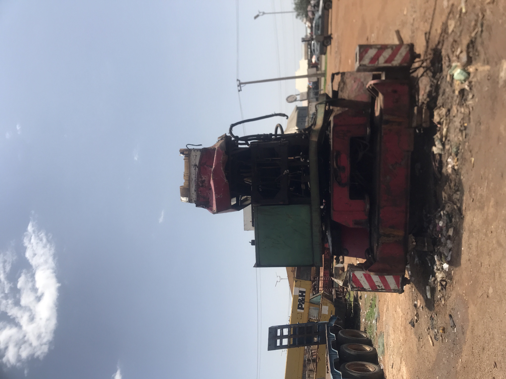 The image shows a large, red and green crane in a cluttered outdoor lot. The crane appears to be old and possibly damaged. In the background, there are other vehicles, including a yellow P&H trailer, and buildings that suggest an industrial or storage area. A power line and utility pole are visible in the distance. The ground is dirt and strewn with debris. The sky is clear with some scattered clouds. The location is identified as Rafin Par, Nigeria.