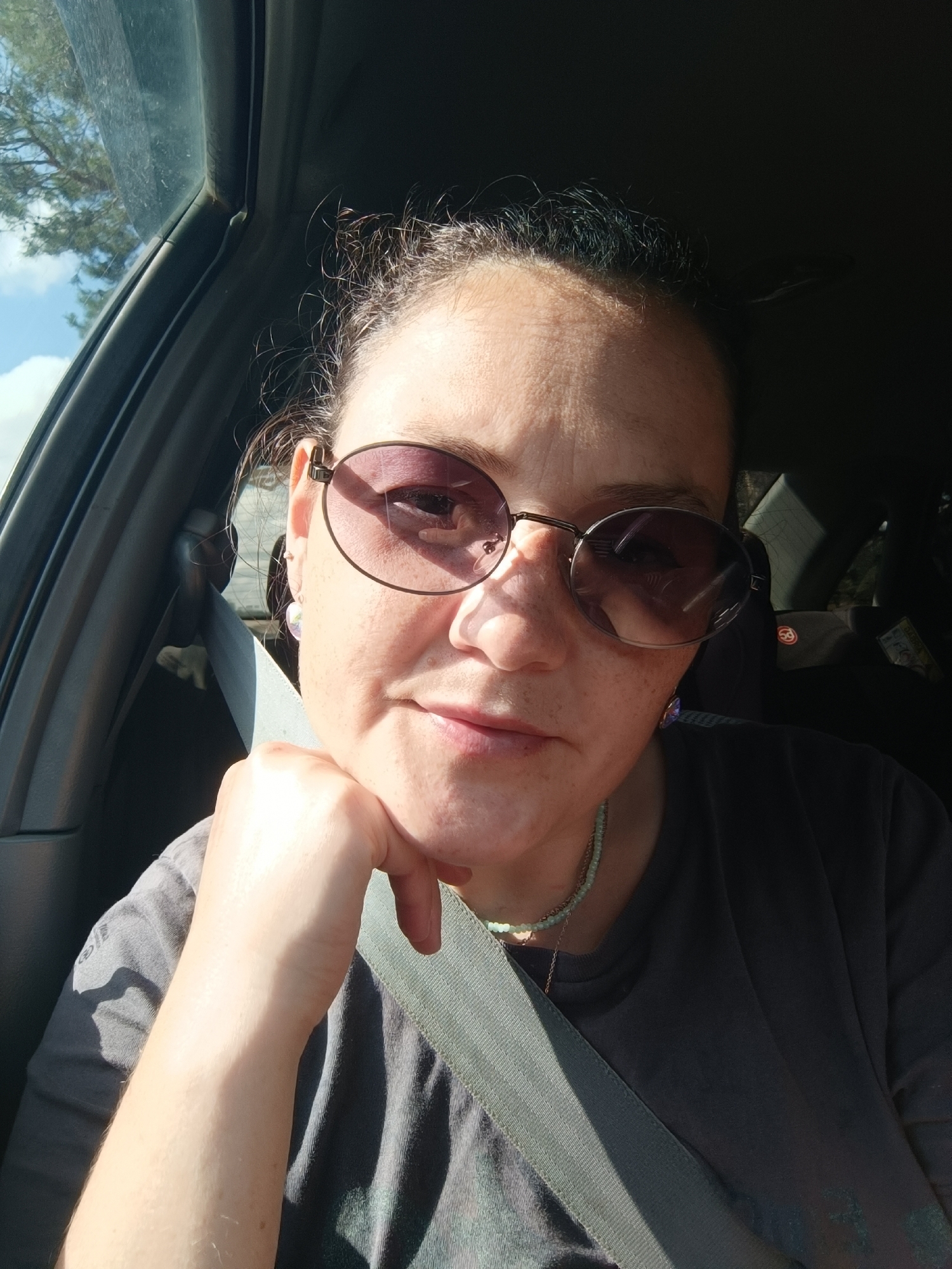 A medium shot photograph shows a woman sitting in a car, looking directly at the camera. The woman has dark hair pulled back and is wearing large, round sunglasses with tinted lenses. She is resting her chin on her left hand, which is supported by her right hand. She is wearing a dark gray t-shirt and a beaded necklace. A seatbelt is visible across her chest. The background shows the interior of a car, with a window on the left revealing trees and a blue sky with clouds. The lighting suggests it is daytime, with sunlight casting shadows. The scene appears to be a selfie taken by the woman. There is no specific indication of the city of Foki, Russia, in the image.