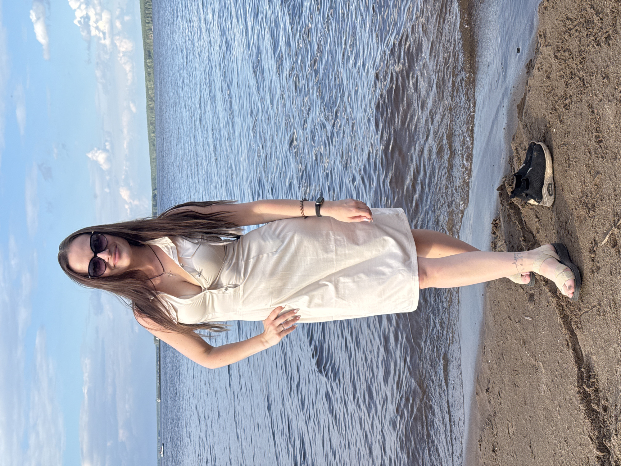 The media file shows a young woman standing on a sandy shore next to a body of water. She is wearing a light-colored dress, sunglasses, and sandals. Her hair is long and dark, and she is smiling at the camera. To her left, a black Adidas sneaker is visible on the sand. The water appears calm, with small ripples on the surface. The sky is blue with scattered clouds, suggesting it is daytime and the weather is pleasant. A thin line of vegetation can be seen in the distance across the water. The image is oriented vertically. There is no visible text in the media file. The setting is likely a beach or lakefront in Chaikovsky, Russia, given the provided context. The woman appears relaxed and happy.