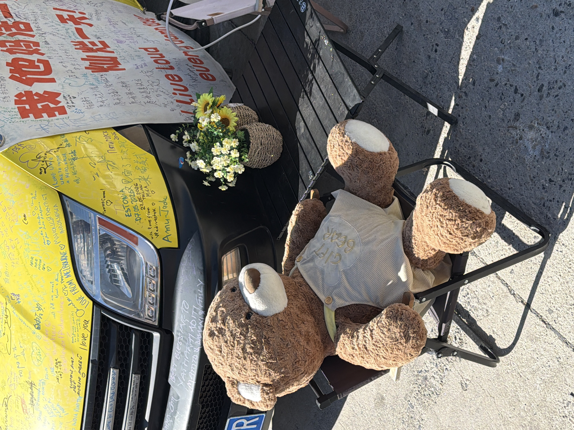 The image captures an outdoor scene, likely during a daytime stop on a long journey, in Gyirong County, China. The primary subjects are the front section of a yellow vehicle and a large brown teddy bear.
The vehicle's hood and front grille are visible, with the yellow surface extensively covered in handwritten messages, signatures, and drawings in various languages. A prominent headlight is also seen. Above the vehicle, two white banners are displayed; one features large red Chinese characters, while the other shows English text, including "Live Loud eventu." Both banners, like the vehicle, are covered with numerous handwritten notes.
Next to the vehicle, a large, fluffy brown teddy bear is seated in a black folding chair. The bear wears a light-colored, possibly striped, vest or shirt with "GIFT BEAR" embroidered on it. Its white muzzle and inner ear areas stand out. Beside the bear, on what appears to be a small black folding table, are two woven baskets holding artificial sunflowers and small white flowers, adding a decorative touch.
The multitude of messages on the vehicle and banners suggests a significant road trip or expedition. Visible text in the OCR includes "INDIA ROAD TRIP," "LOVE FROM MELBOURNE," "UAE TO INDIA," and mentions of "Estonia," "Tallinn," "Vladivostok," "Tibet," "Piatigorsk," "Indonesia," indicating an international journey. Messages are written in English, Chinese, and other languages like Hindi/Devanagari script, expressing well wishes and personal notes (e.g., "I love you," "BEST BLESSING TO"). Dates like "07.05.2026" and "27.05.2026" are also present, possibly indicating future or past milestones of the trip. The bright sunlight suggests clear weather. The teddy bear appears to be a mascot or companion for this extensive adventure.