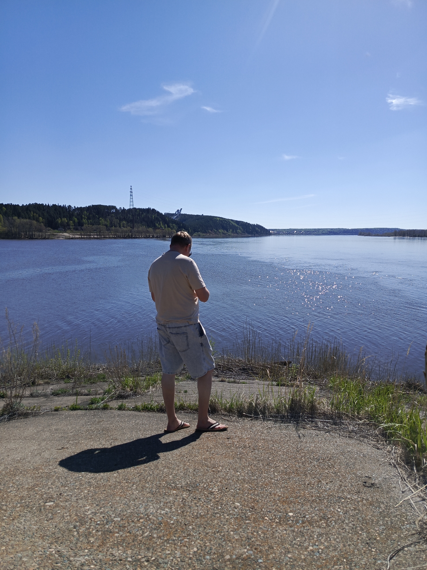 A medium-full shot depicts a man standing with his back to the viewer on a concrete embankment, overlooking a wide river on a sunny day.
The man is fair-skinned, wearing a short-sleeved tan t-shirt, light blue denim shorts, and sandals. His shadow is clearly cast on the concrete surface.
Behind him is a wide, blue river reflecting sunlight. Across the river, there is a forested area. A tall transmission tower and what appears to be a large crane or bridge construction are visible against the skyline.
The location is likely near the Kama River in the city of Chaikovsky, Perm Krai, Russia, based on landmarks present in the photo. Tall grasses and weeds grow along the edge of the embankment. The sky is clear with a few sparse clouds.
