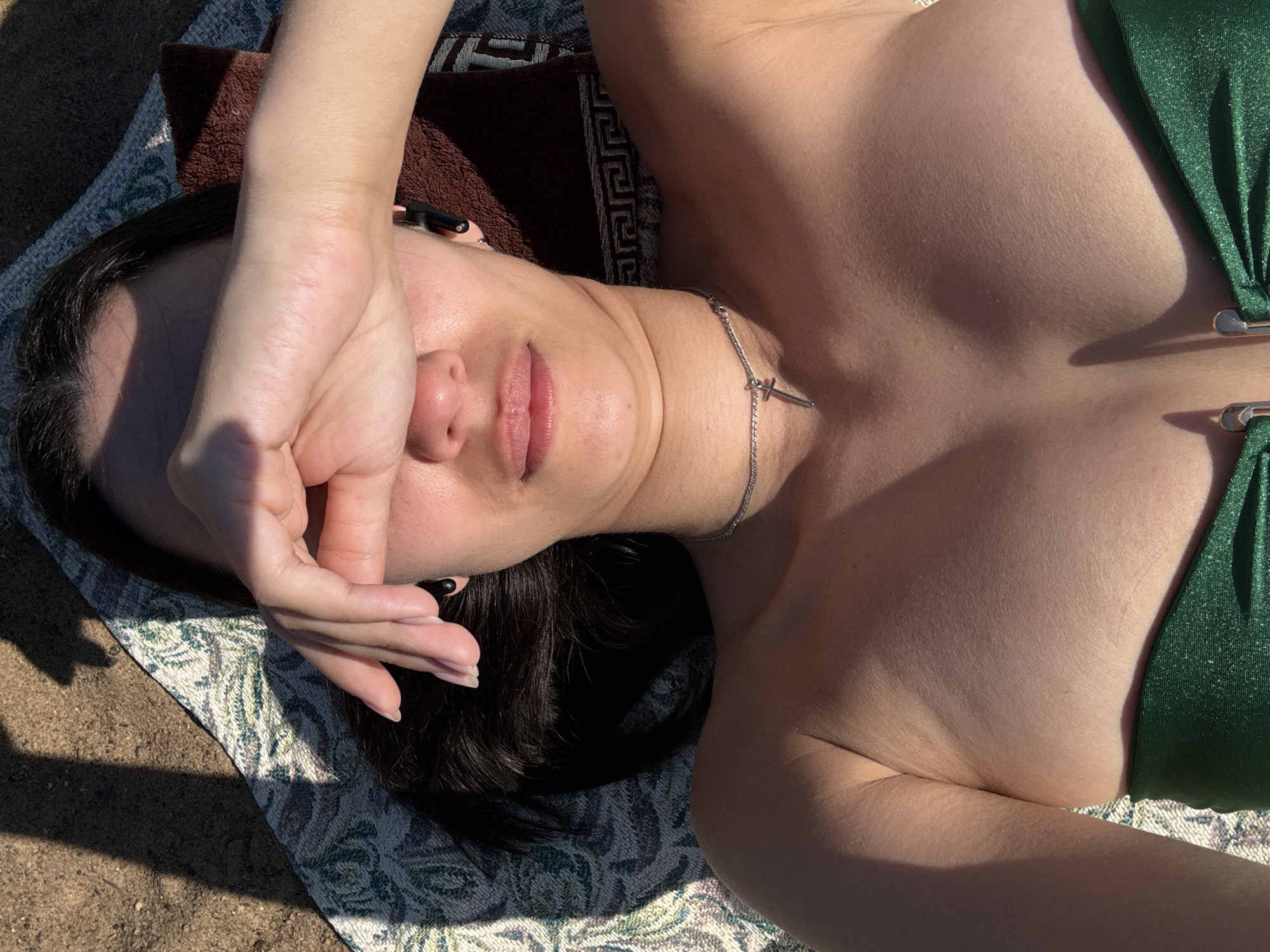 The image shows a woman lying on a patterned blanket on a sandy surface, likely a beach. She is wearing a green bikini top and a silver necklace with a cross pendant. Her dark hair is partially spread on a brown towel underneath her head. She is covering her face with her hand, shielding her eyes from the sun. The location is potentially a beach in or around the city of Chaikovsky, Russia. The lighting suggests it is daytime. There are no other visible people or interactions in the scene.