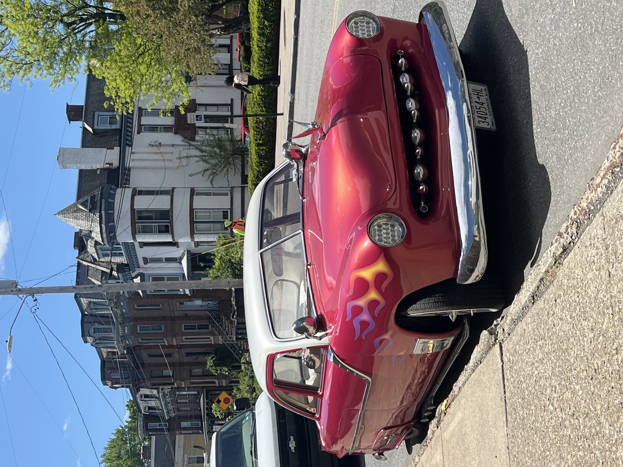 The image captures a bright, sunny day scene in what appears to be a residential area of the City of Newburgh, United States of America. The foreground is dominated by a red and white vintage car parked along the side of a street. The car has chrome accents and flame decals on its front fender. It also has a New York license plate that reads "34054-HL HISTORICAL". The background showcases a row of buildings, including a distinctive three-story brick building with white trim. There is a person in dark clothing walking along the sidewalk in front of the buildings, appearing small in comparison to the buildings. A green hedge is visible, adding a touch of natural greenery to the urban landscape. The sky is clear, and the shadows are dark and defined, which indicate that it is midday. A utility pole and a yellow street sign are visible on the left side.