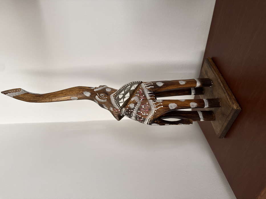The image displays a handcrafted wooden elephant sculpture positioned indoors. The specific city location within AMCO, Area Metropolitana Centro Occidente, Colombia, is not discernible from the visual information provided. The sculpture, crafted from dark brown wood, features an elongated, stylized elephant standing with its trunk raised and curved upwards. It is adorned with white painted patterns, including dots on its legs and trunk, stripes on its lower legs, and a geometric motif on its back/saddle area. Additional swirling patterns in white and reddish-maroon are present on the body. The elephant stands on a flat, rectangular base of lighter brown wood, exhibiting minor wear. This base rests on a polished, dark reddish-brown wooden surface, likely a table or shelf, which occupies the lower portion of the frame. The background consists of a plain white wall, forming an interior corner on the left side of the image. No people, actions, or interactions are visible.