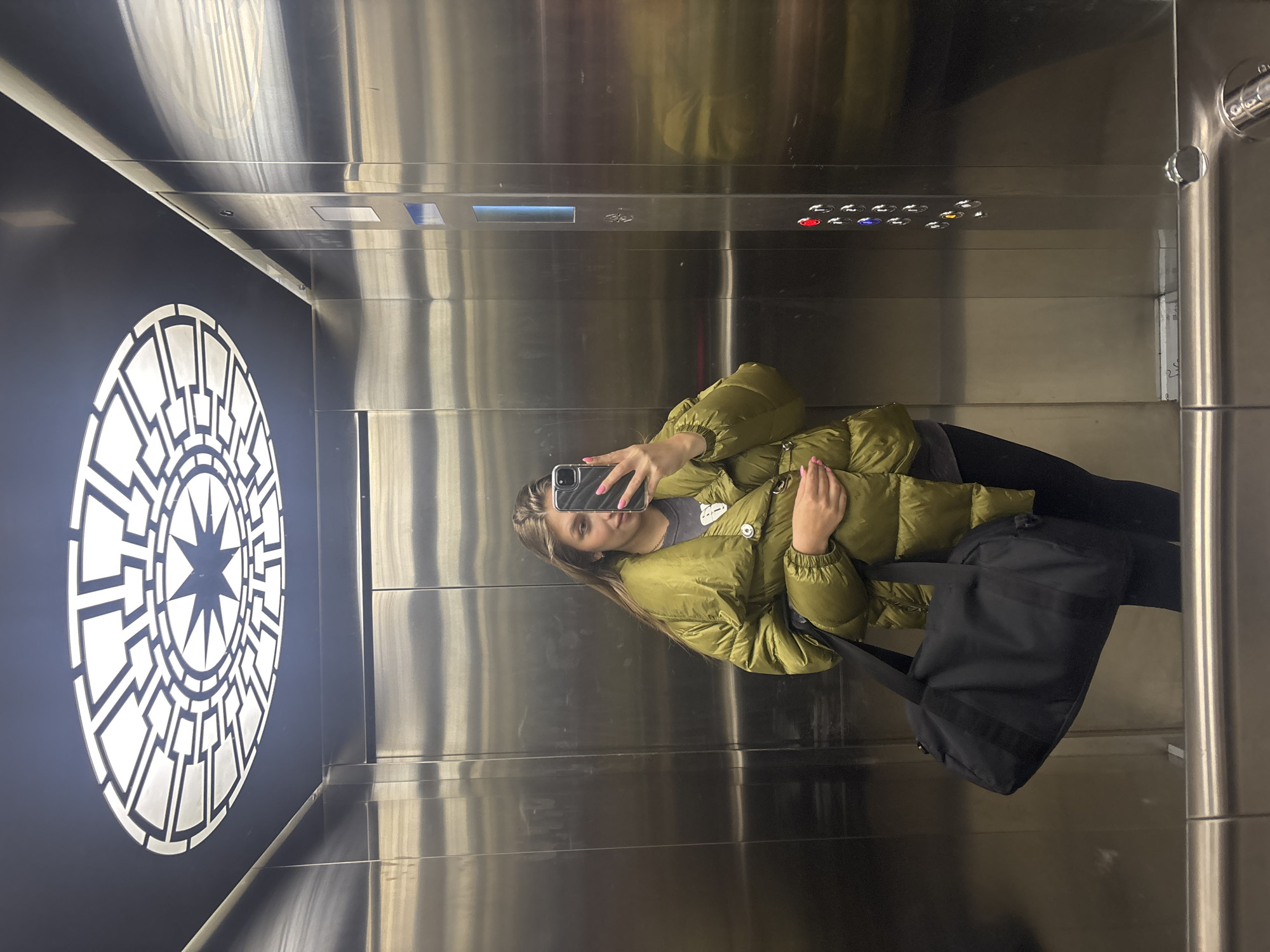 A woman is taking a selfie in an elevator. She is wearing a green puffer jacket, black leggings, and carrying a black tote bag. The elevator has stainless steel walls, a decorative circular design on one wall, and buttons for different floors. The city is Kharkiv, Ukraine.