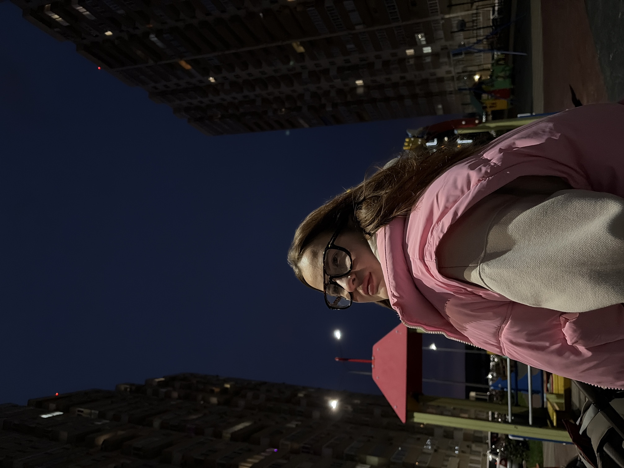 An image, presented rotated 90 degrees clockwise, captures a young woman at night or late twilight in an urban setting in Krasnodar, Russia. She is shown from a low angle, wearing a pink puffer vest over a light-colored top, and black-rimmed glasses. Her dark hair falls around her shoulders, and she looks towards the camera with a neutral expression.
The background features two large, dark residential or commercial buildings, positioned on either side of the frame, with numerous windows illuminated, suggesting an active urban environment. The sky is a deep, dark blue, indicating nightfall. Several bright, circular light sources, possibly the moon or prominent streetlights, are visible in the dark sky. To the lower right, glimpses of a colorful structure, likely part of a children's playground, can be seen with elements in red, blue, and yellow. The overall lighting is dim, relying on artificial light from the buildings and some ambient sources. No visible text is present in the image.