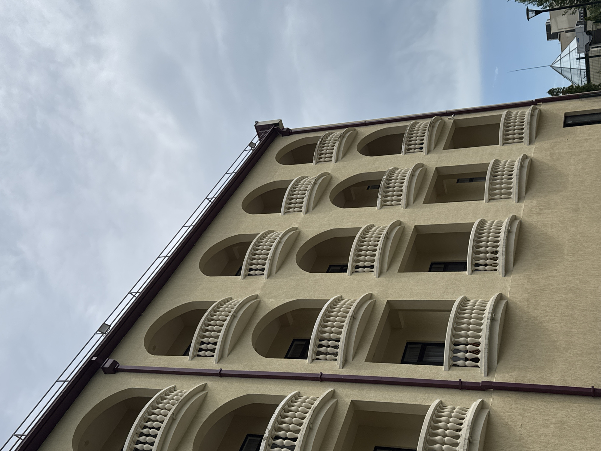 A low-angle, full shot captures a tan, stucco building with multiple levels of arched balconies. Each balcony features a white, ornate railing. The building has a dark red trim along the roofline, which is equipped with a metal railing and a floodlight. A dark red downspout runs vertically along the left side of the building. The sky above is overcast and a light gray, with patches of blue visible. In the background, to the right of the building, a glass pyramid structure and a lamppost are visible.