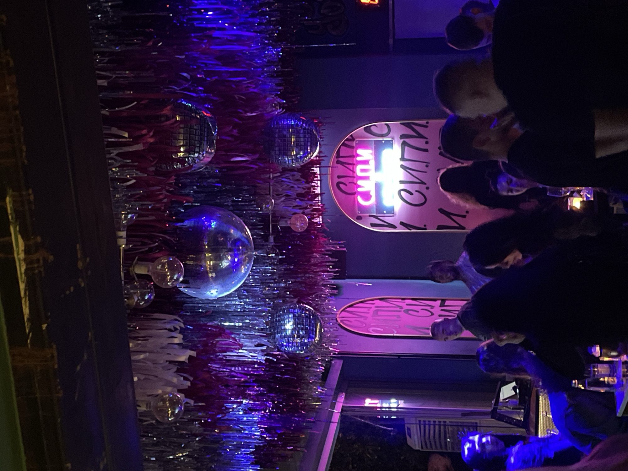 The image depicts a dimly lit bar or club, likely located in Plovdiv, Bulgaria. The decorations are notable, featuring multiple disco balls of varying sizes hung against a backdrop of silver and red tinsel. There is a sign with Cyrillic text, glowing in neon pink, which adds to the ambiance. Patrons are present, standing and interacting, but are somewhat obscured by the low lighting and focus on the decor. The general mood is that of a nightspot with a festive or celebratory atmosphere.