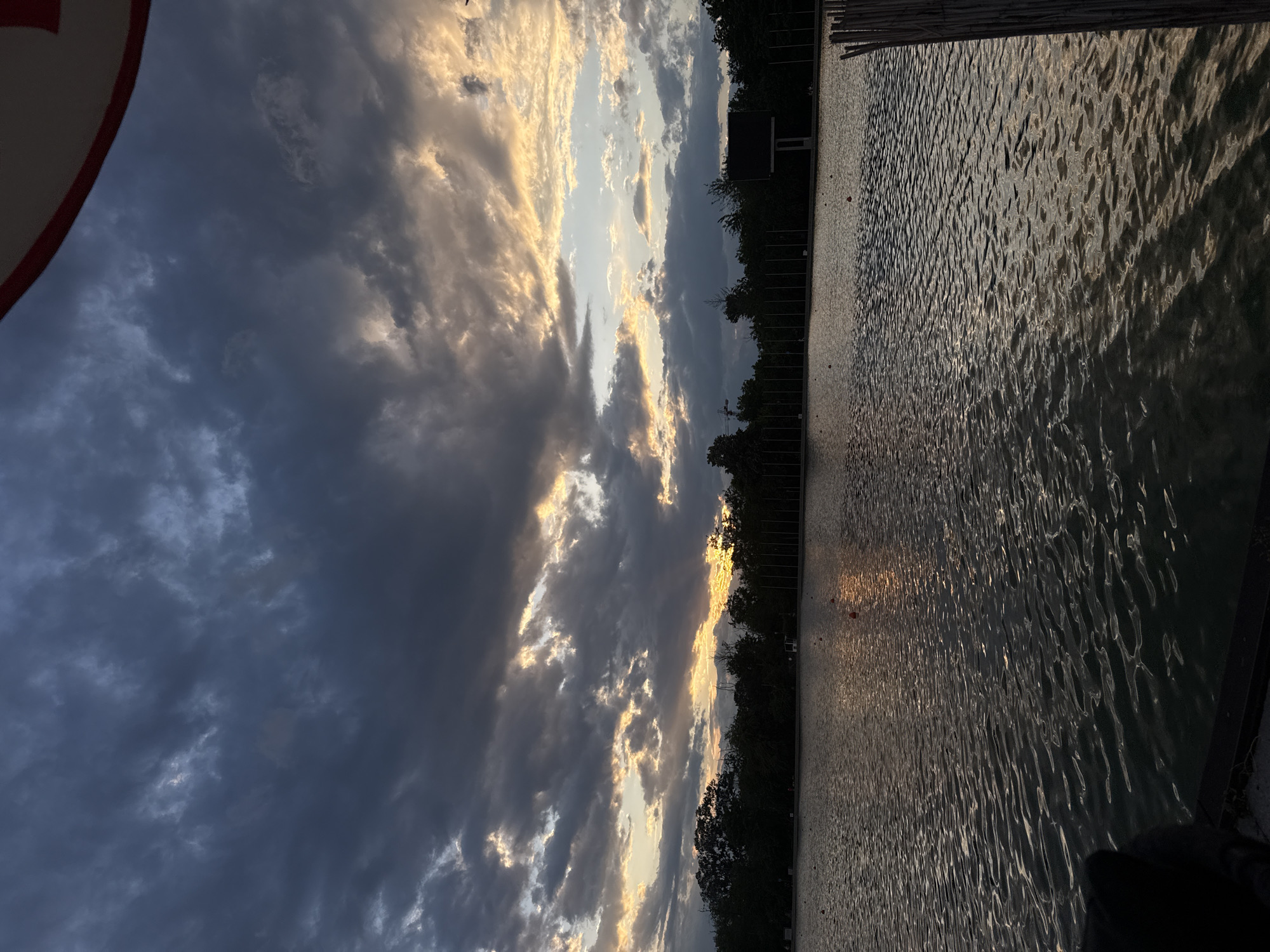 The image displays a serene landscape scene at either sunrise or sunset, centered on a large body of water under a dramatic sky. The perspective appears to be from a low vantage point, likely on or adjacent to a pier or shoreline.
The sky, occupying the upper half of the frame, is filled with a mixture of heavy, dark grey clouds and bright, golden light. Sunlight, presumably from the low sun, breaks through gaps in the clouds, creating luminous streaks that cast a warm glow on the cloud edges, while other areas remain in shadow.
Below the horizon, a calm body of water stretches out, its surface covered in countless small ripples. These ripples reflect the golden light from the sky, creating shimmering, dappled patterns across the water. The reflections suggest the sun is low, just above or behind the distant tree line.
Along the far side of the water, a dense line of dark, silhouetted trees forms the shoreline. Above these trees, a distinct dark, rectangular structure, possibly a building or part of a facility, is visible.
In the immediate foreground on the right, a section of a dark wooden structure, likely a pier or dock, extends into the scene. In the extreme upper-left corner, a partial view of a red and white circular object, possibly a buoy or a safety sign, is present. No people are visible, and the overall atmosphere is one of tranquility and natural beauty. The setting is in Plovdiv, Bulgaria.