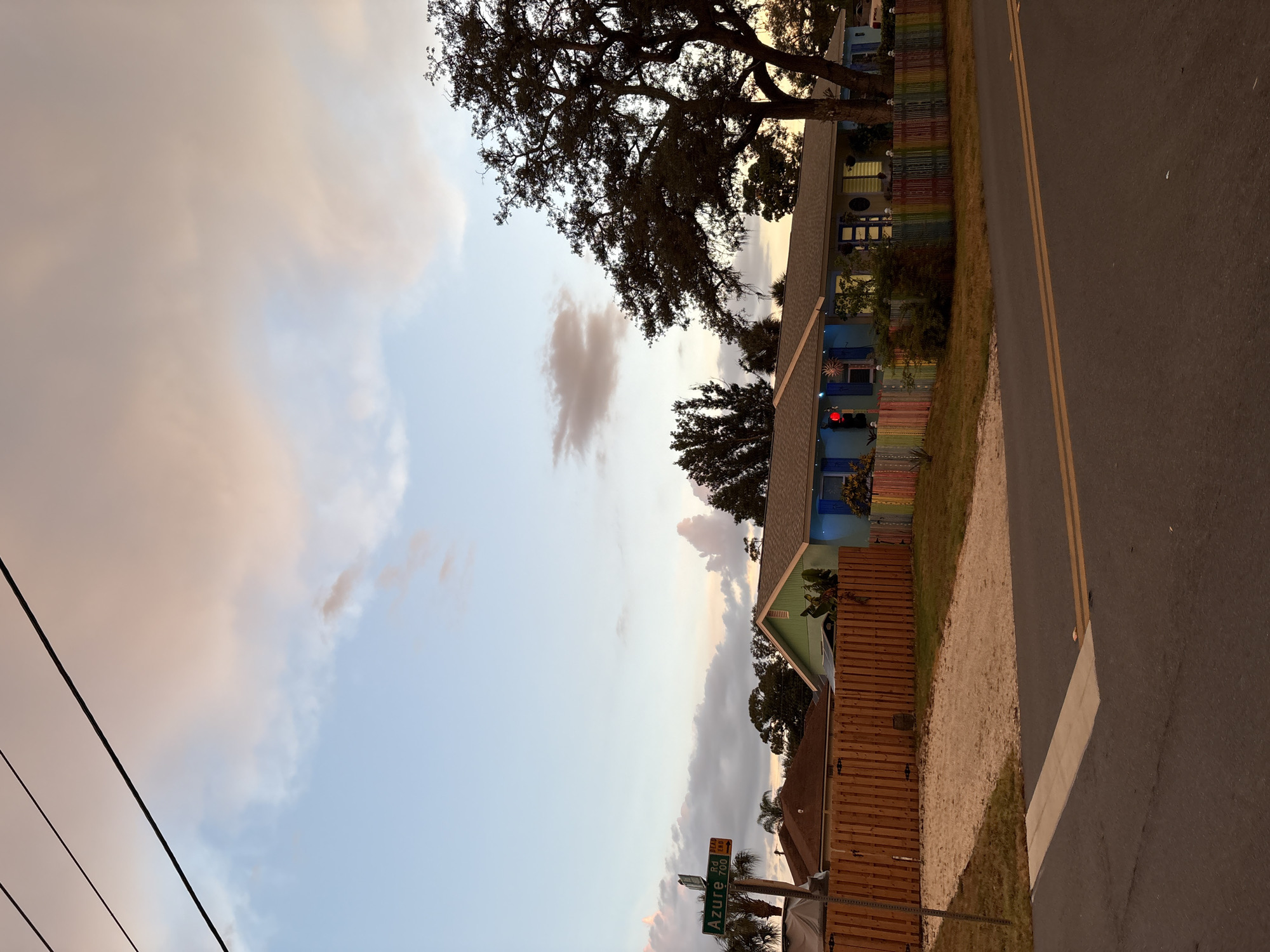 The image captures a residential street scene in South Venice, Florida, during twilight. The sky displays a gradient of pale blue and soft pink clouds, illuminated by the setting sun. Overhead, dark power lines stretch across the frame. A street sign indicates "Azure Rd 700" with "TEA END" and a directional arrow. In the foreground, asphalt pavement with double yellow lines forms a road, with a concrete crosswalk visible.
To the right of the road, a property is bordered by a wooden fence. Beyond the fence, a low-slung house features a vibrant, multicolored fence in front, adorned with various colors such as red, orange, yellow, green, blue, and purple. The house itself is painted in a cool tone, possibly blue, with blue shutters and a red traffic light signal prominently displayed on the left side of the property. A large, mature tree with dense foliage stands to the right of the house, casting a silhouette against the sky. In the background, other houses with brown roofs and wooden fences are visible, along with palm trees. The overall impression is that of a suburban neighborhood at dusk.