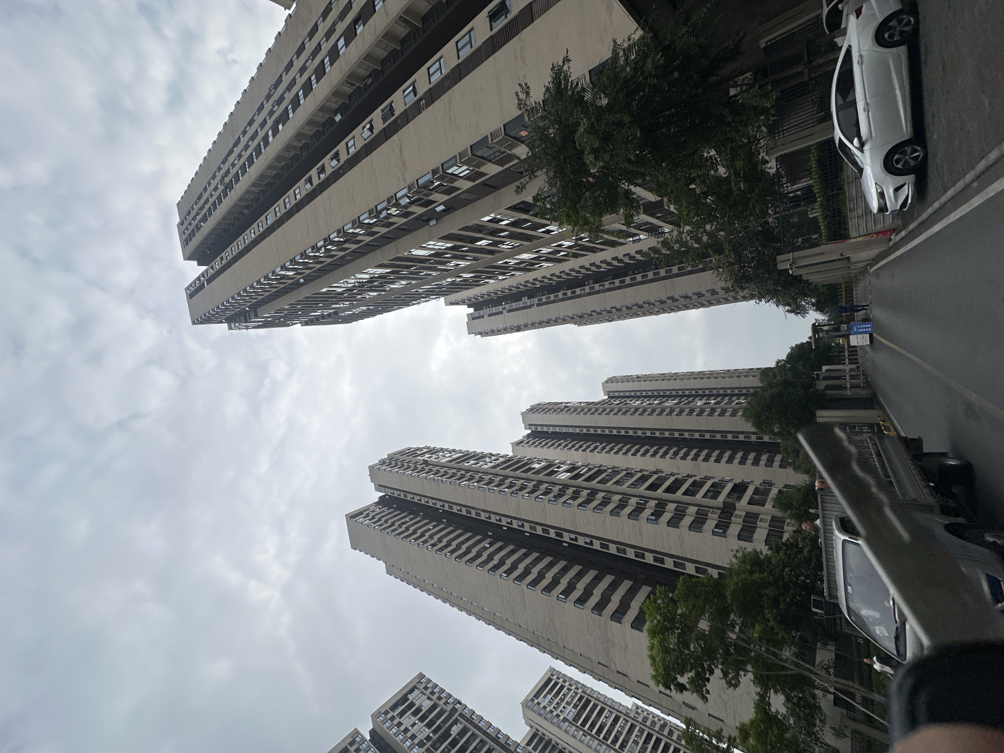 The image, taken in Chongqing High-tech Zone, China, shows a low-angle view of tall, densely populated residential buildings under a cloudy sky. Several high-rise apartments dominate the scene, their facades marked by repetitive window patterns and horizontal architectural bands. Trees are interspersed among the buildings, partially obscuring some structures. A paved road runs alongside the buildings, and a white car is parked near the buildings to the right. Another car or van is visible on the right side of the image.