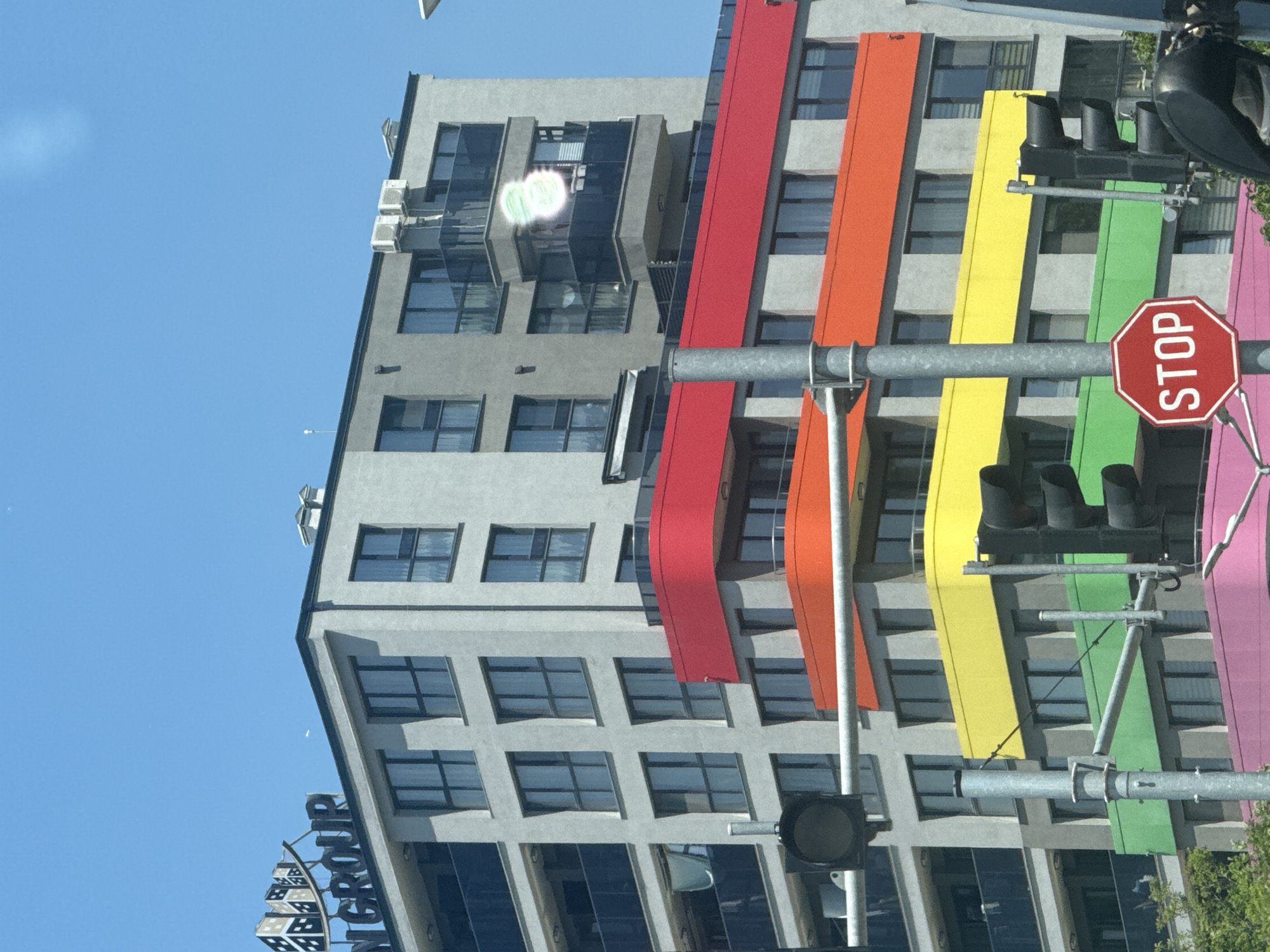 This is a medium-shot, eye-level, vertical, outdoor, daytime photograph taken in Varna, Bulgaria. The image features a modern, multi-story building with a gray facade. The building is adorned with horizontal bands of vibrant colors, including red, orange, yellow, green, and pink. Several windows with dark frames are visible on the gray sections of the building, and some balconies with glass railings are also present.
In the foreground, traffic lights are mounted on metal poles. One traffic light is black with three lenses. To the left of the center, another black traffic light is partially visible. A red octagonal stop sign with white lettering is positioned in the lower right quadrant of the image.
The top of the building is topped with a logo that appears to say "N GROUP" in dark letters, with stylized building graphics above it. In the upper right corner of the building, there are visible air conditioning units. A faint, light green circular anomaly is present on one of the balconies. The sky above is clear and blue.